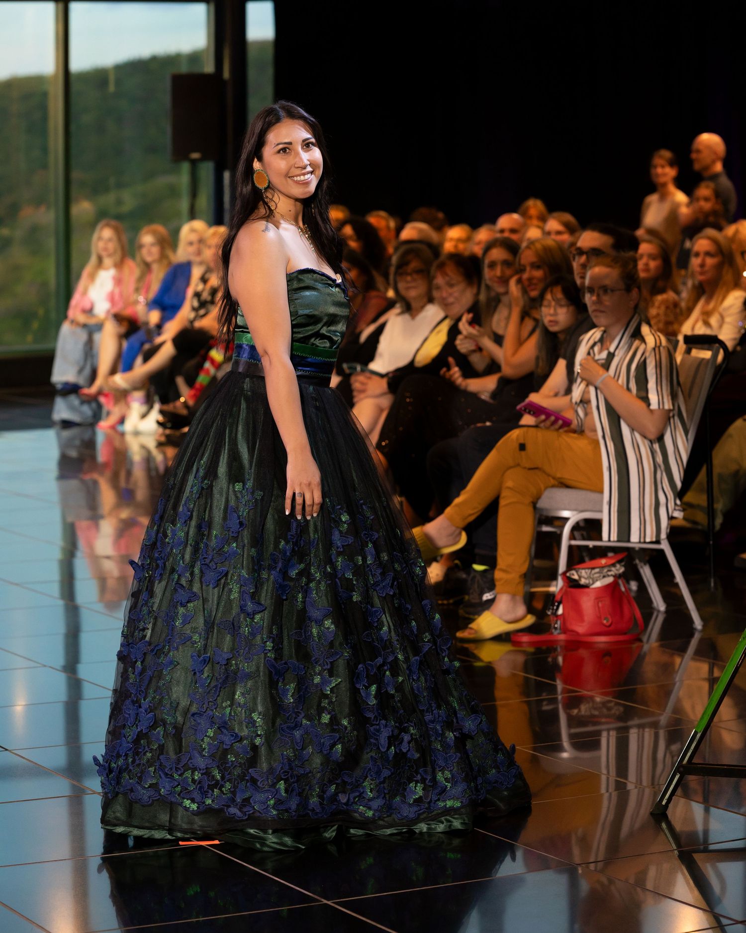 Model wearing an elegant black and navy floor-length gown walks the runway at a fashion show with audience in background.