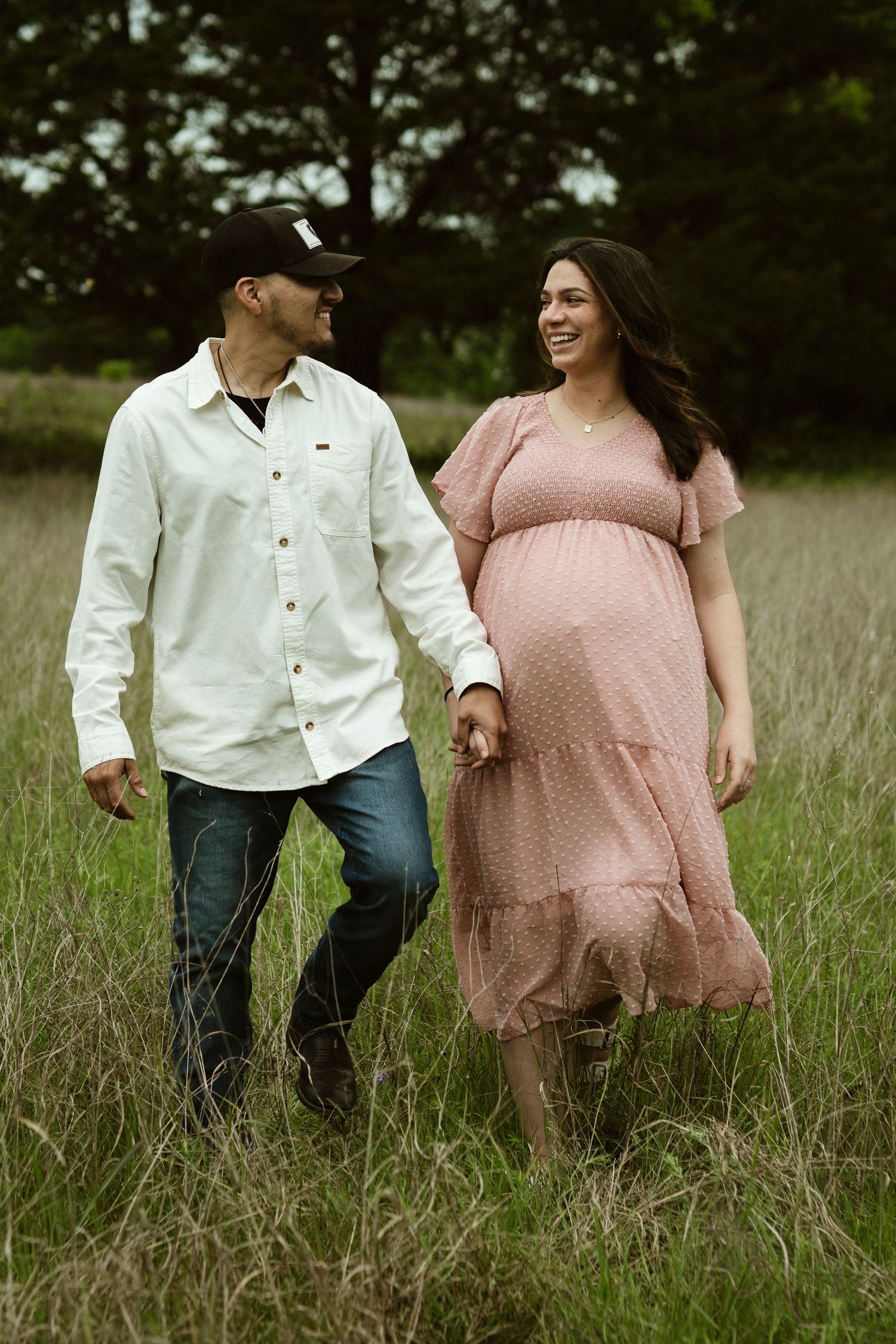 A couple walking through a field of tall grass, with the woman wearing a pink maternity dress.