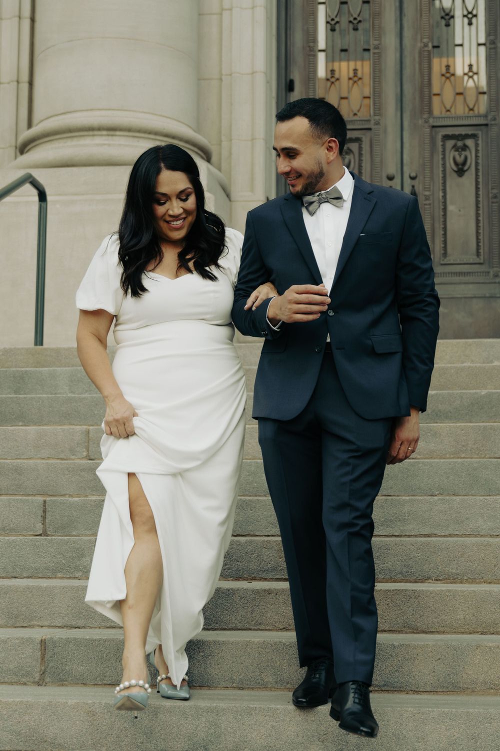 A couple descending stone steps in formal attire, sharing a joyful moment outside a historic building.