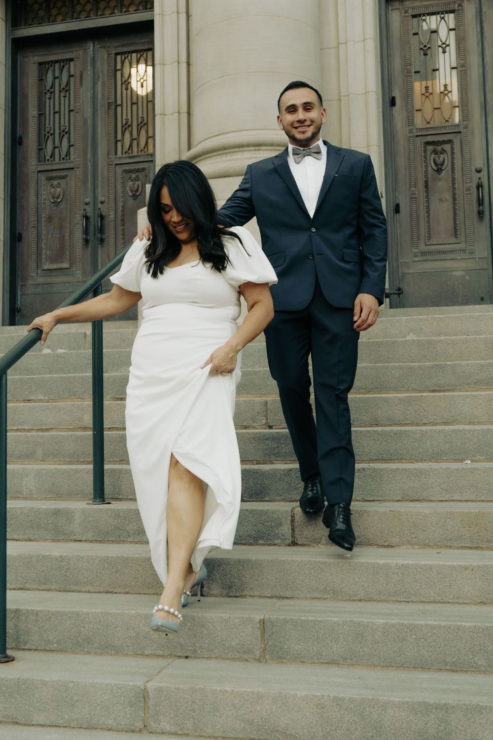 A couple walking down stone steps, with elegant formal wear against classical architecture.