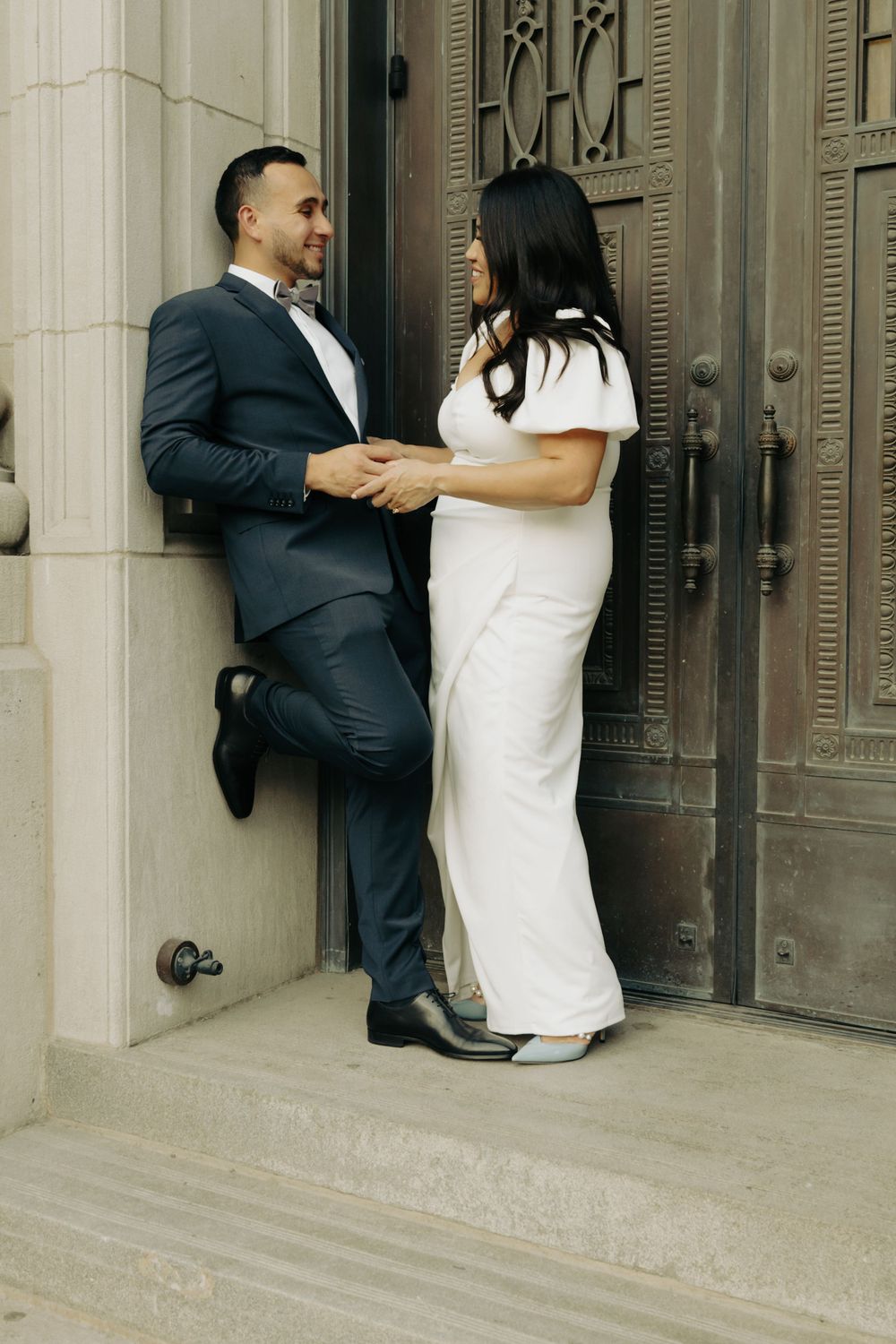 A playful couple in formal attire posing against an ornate door at a historic building.