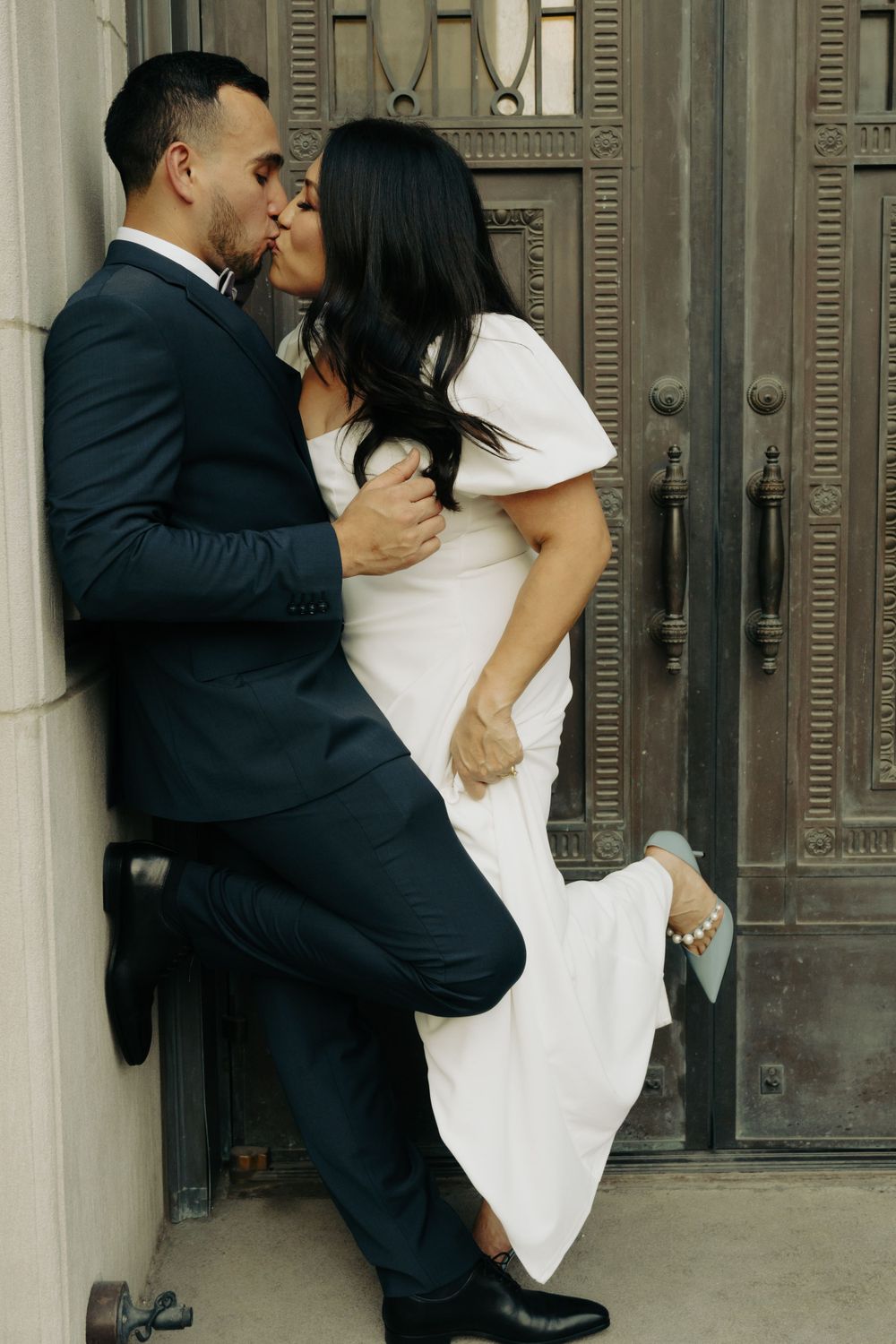 A romantic couple sharing a passionate kiss against an ornate door, with the woman in white and man in dark suit.