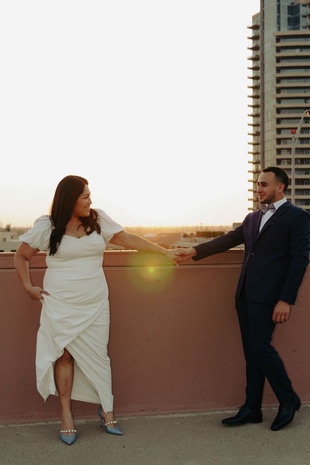 A couple in formal attire dancing playfully on a rooftop at sunset with city buildings in background.