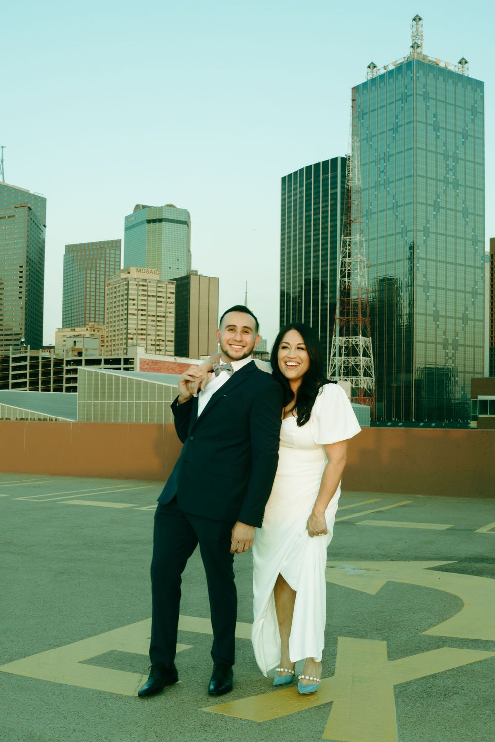 A couple in formal wear posing on a rooftop with downtown Dallas skyline in the background.