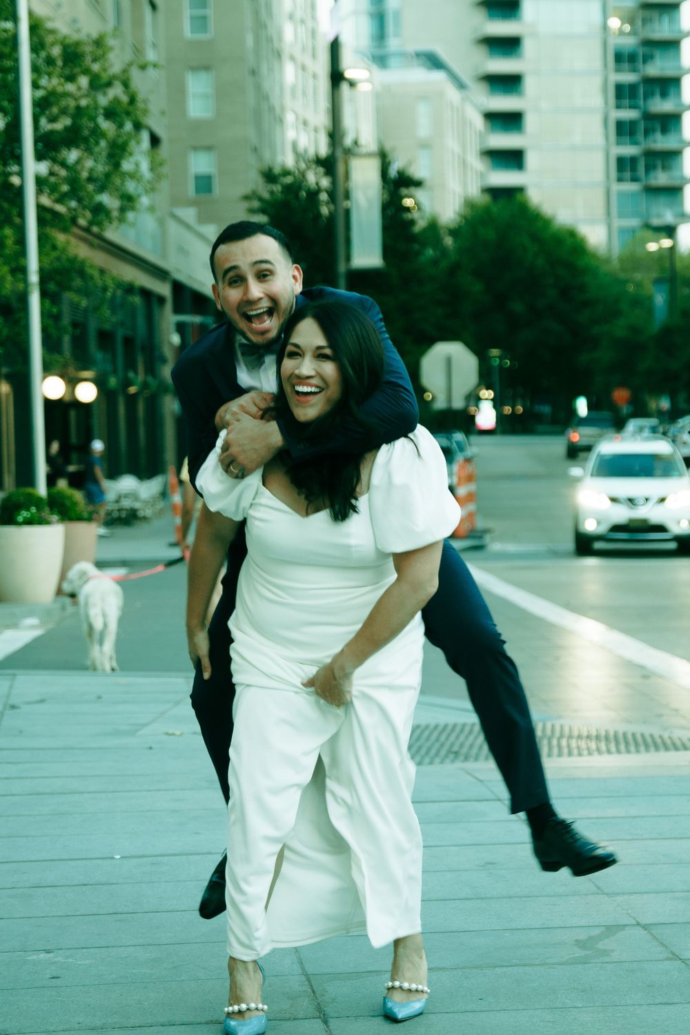 A playful couple sharing a fun moment on an urban street with city buildings in background.