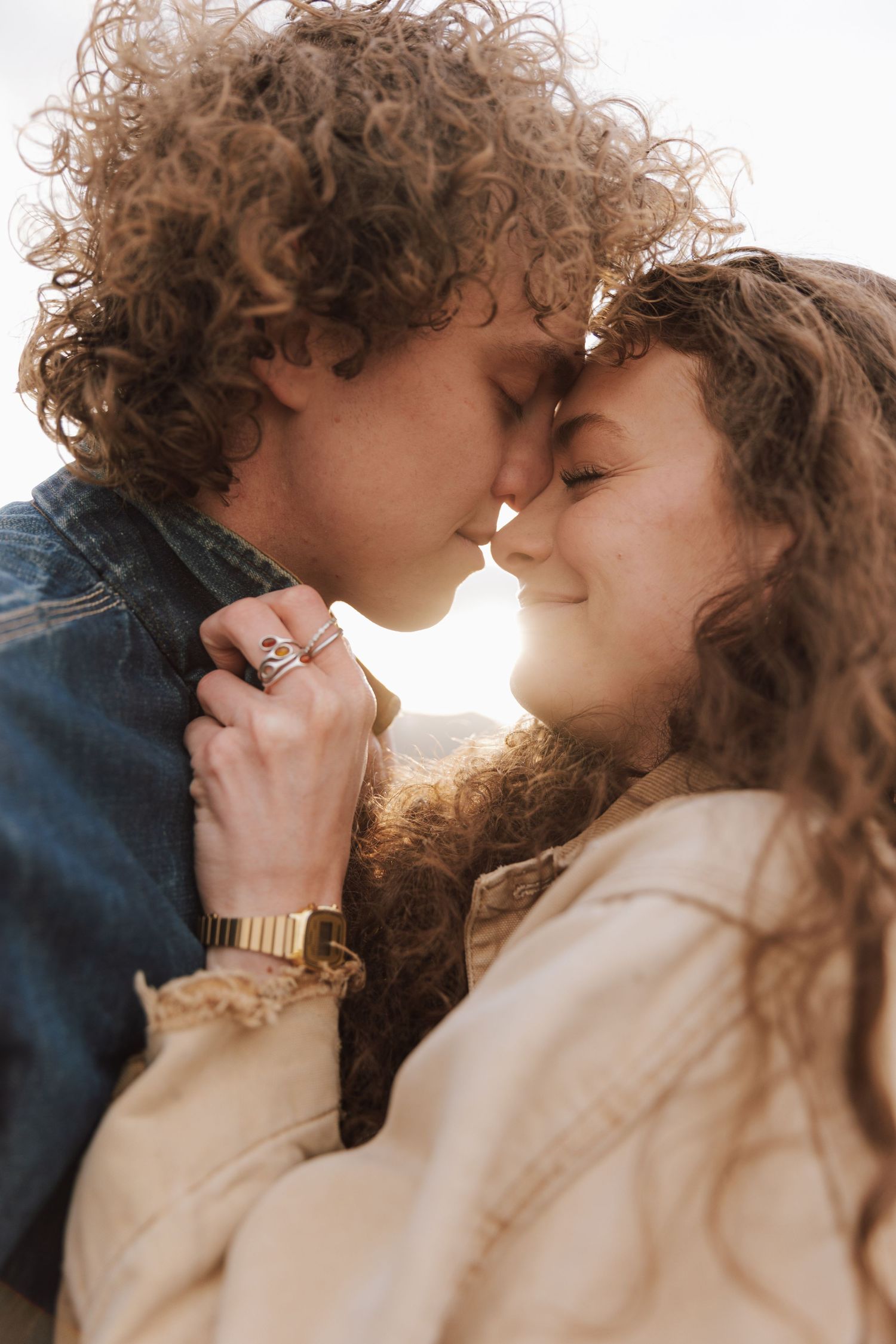 A romantic couple with curly hair embraces outdoors at sunset, both wearing casual clothing in warm lighting.