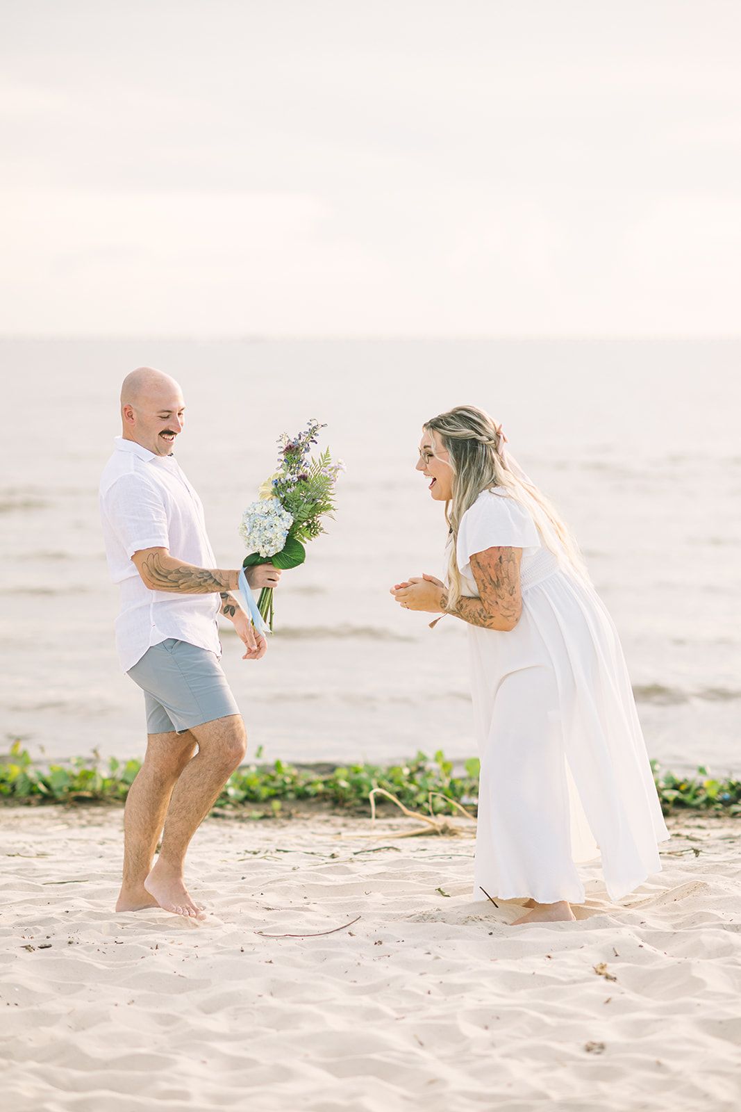 Couple in white and beige outfits playfully interacting on a sandy beach.
