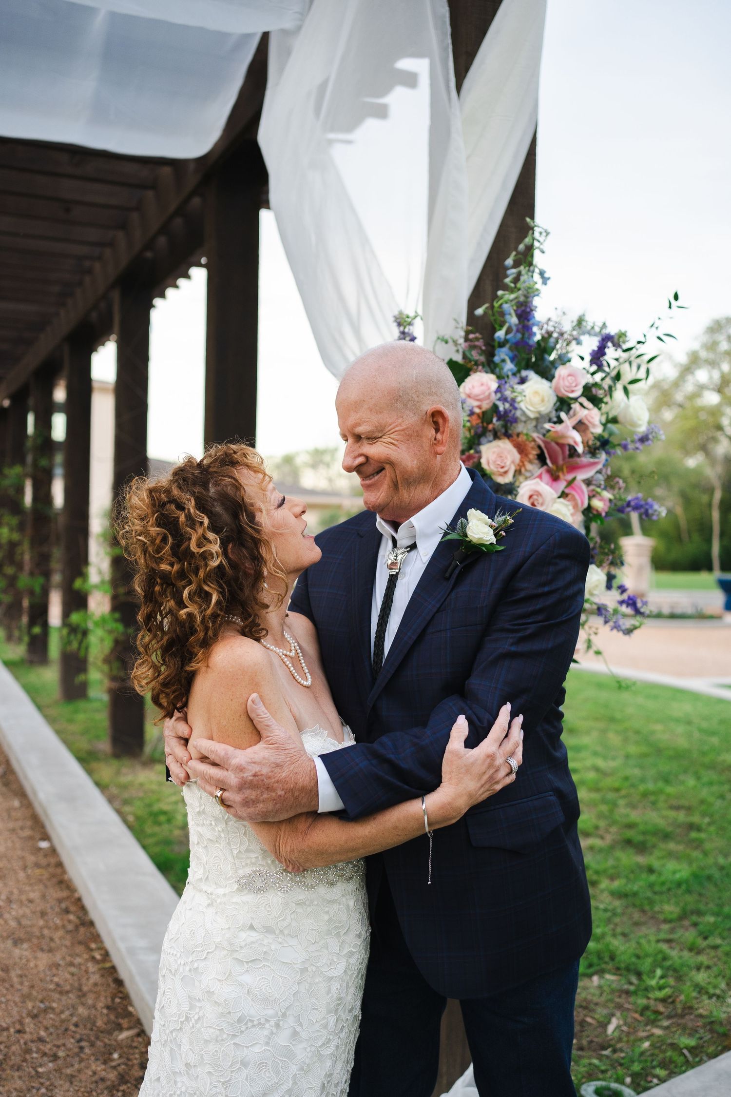 A newlywed couple embraces and smiles at each other under an outdoor chuppah decorated with floral arrangements and flowing white fabric.