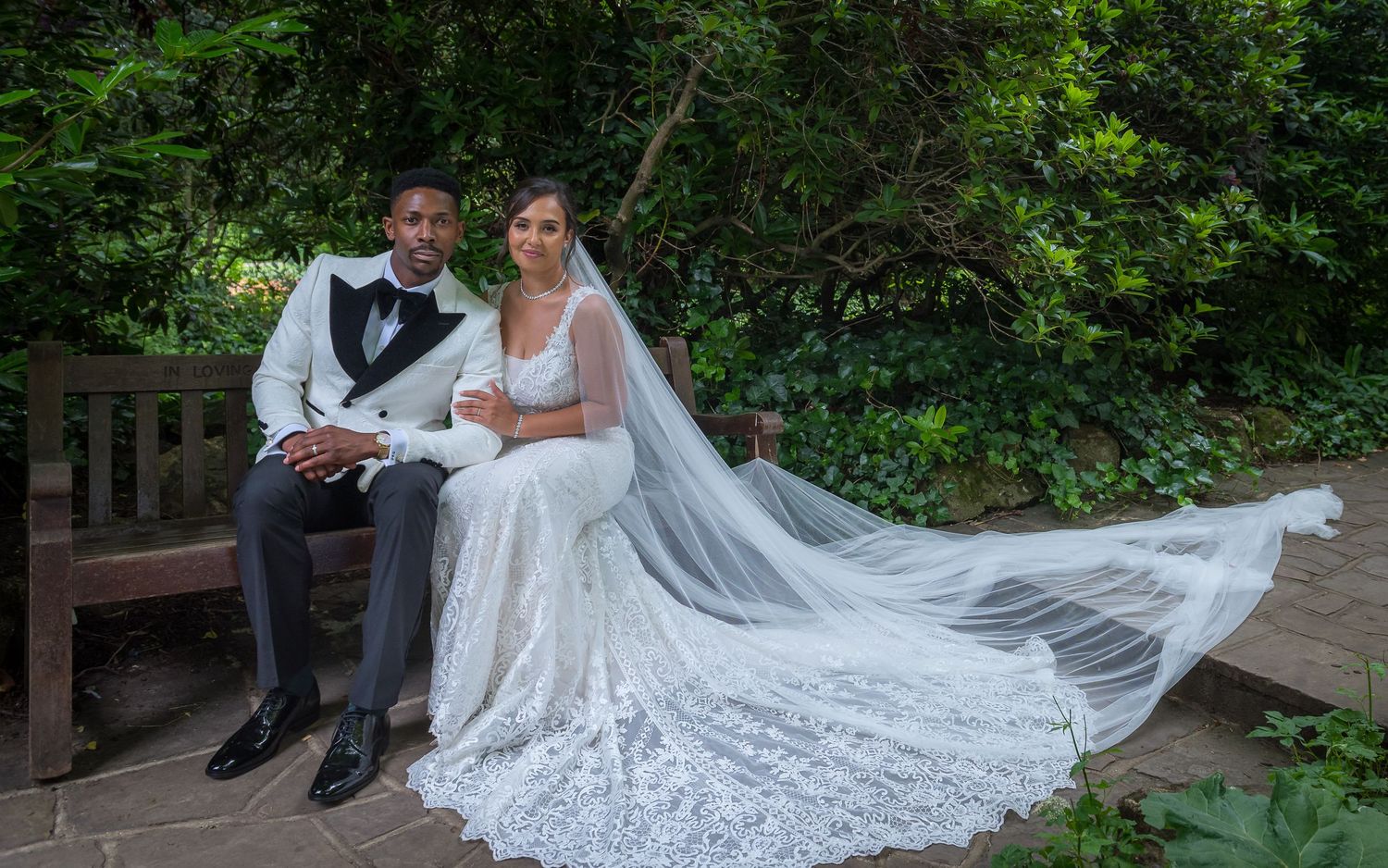 A wedding portrait on a wooden bench with a flowing lace veil in a garden setting.