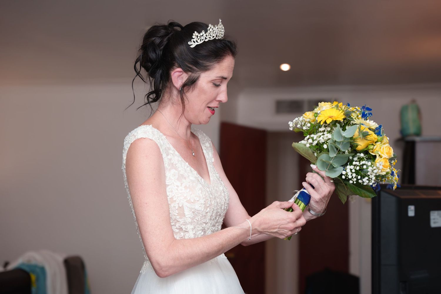 A bride in a lace dress examines a yellow flower bouquet indoors.