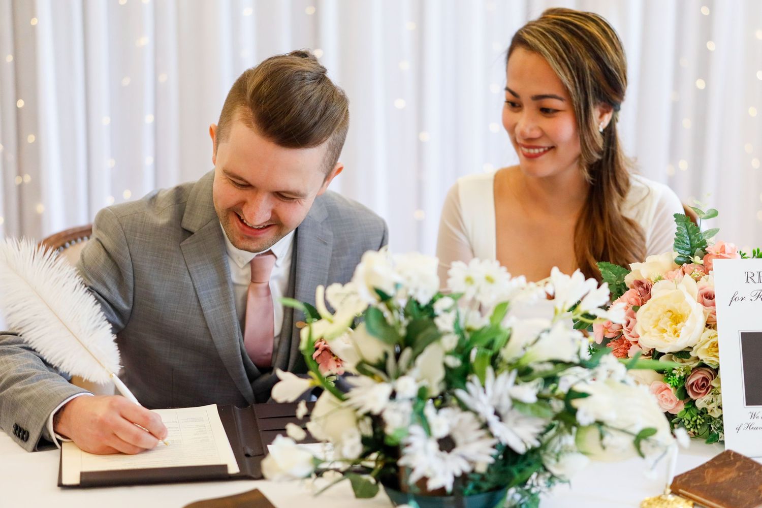 Wedding party signing documents at a table decorated with white floral centerpieces.