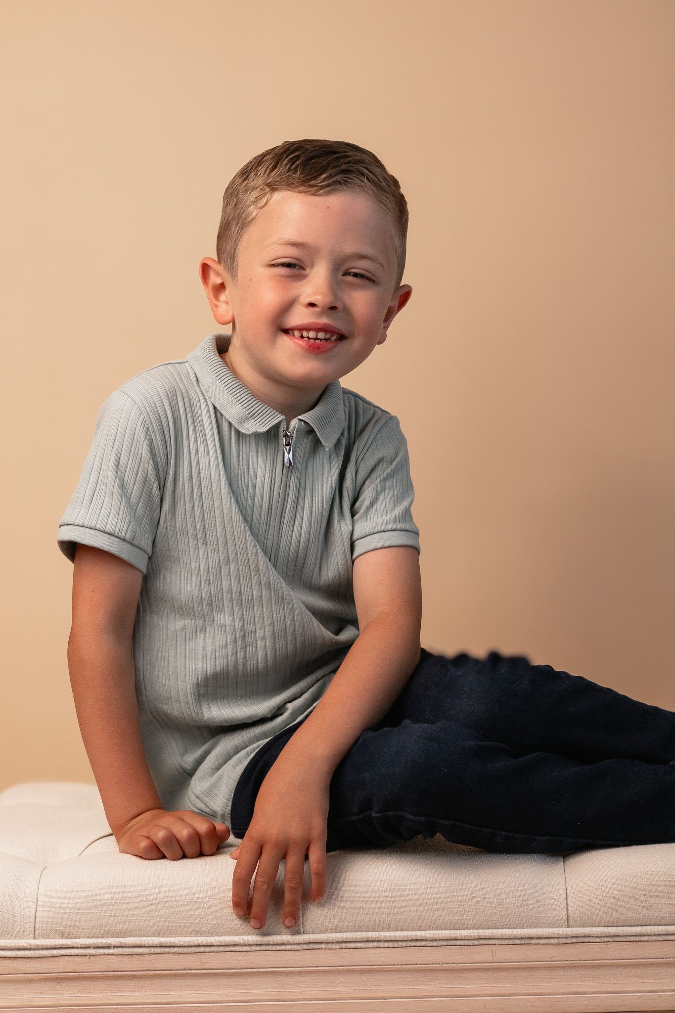 A casual portrait of a child relaxing on a white bench while wearing a grey polo shirt.