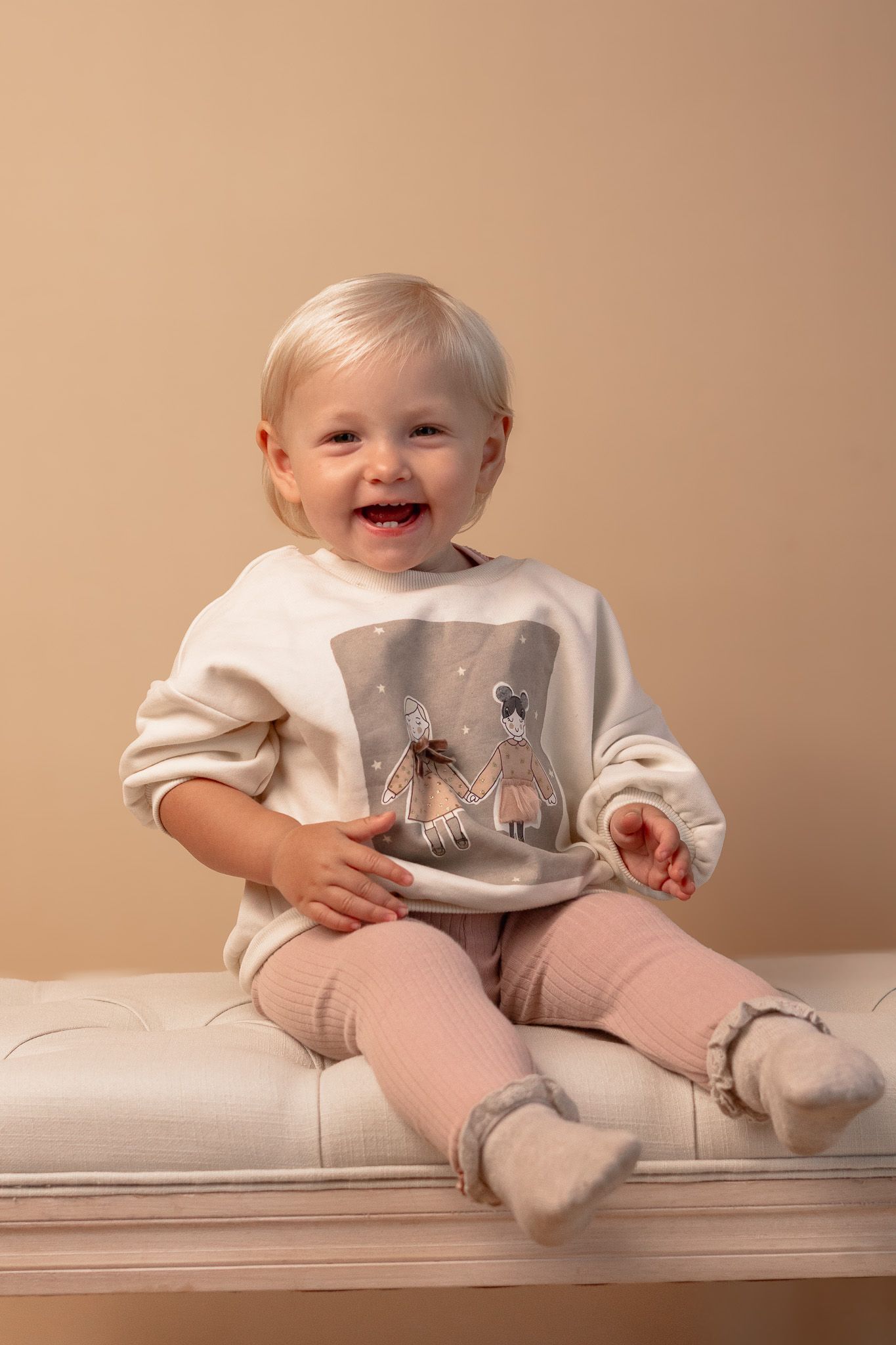 A cheerful toddler in a cream sweatshirt and pink leggings sits on white furniture.