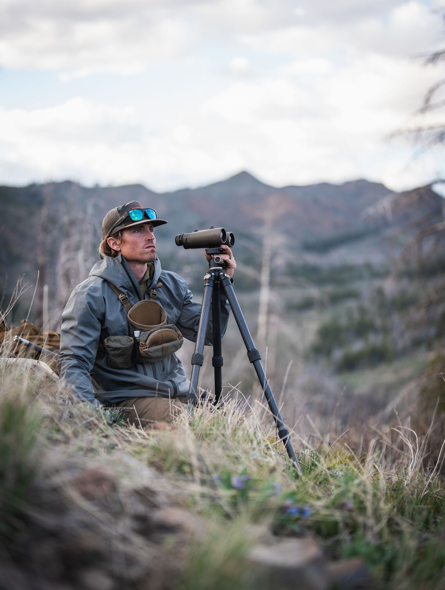 Hunter using spotting scope to scout mountainous terrain during cloudy conditions.
