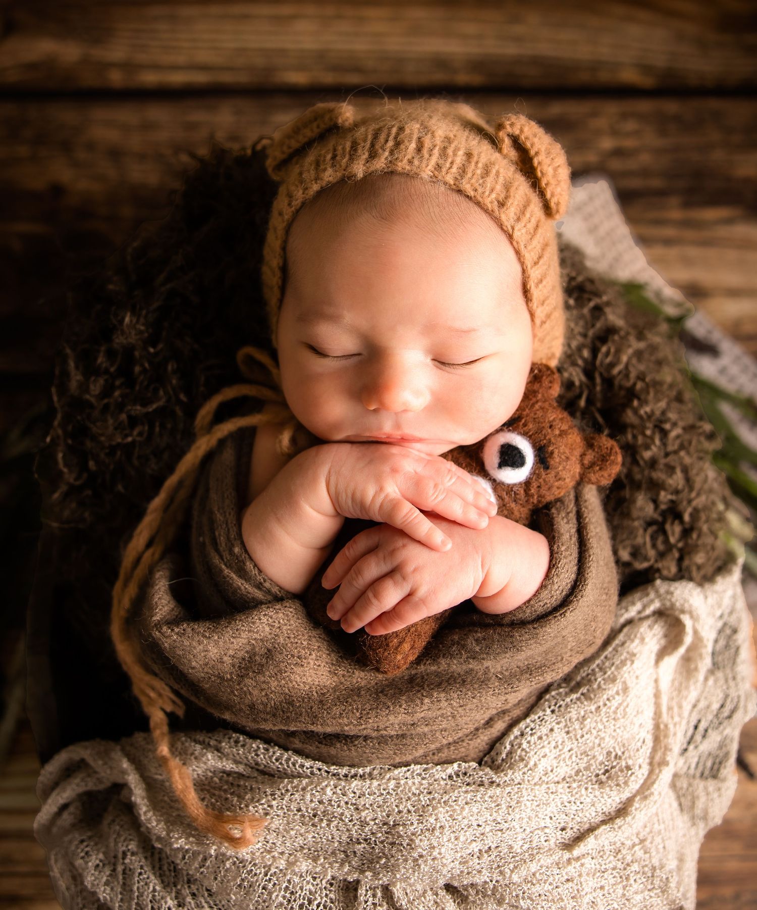 A sleeping newborn wrapped in a brown knit blanket on a rustic wooden backdrop with soft lighting.