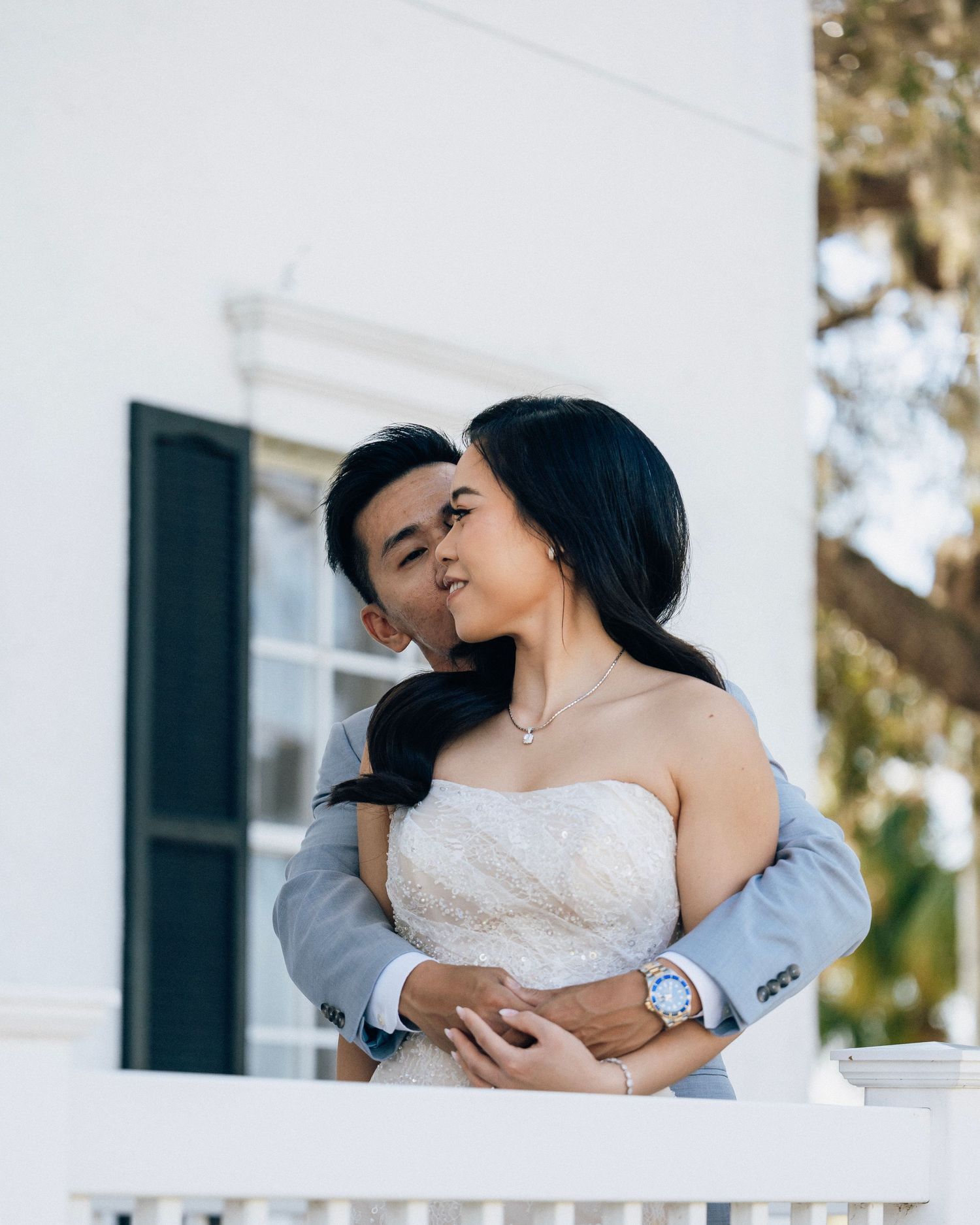 Groom kisses bride on the cheek as she smiles on a white porch balcony, wearing a strapless lace wedding gown.