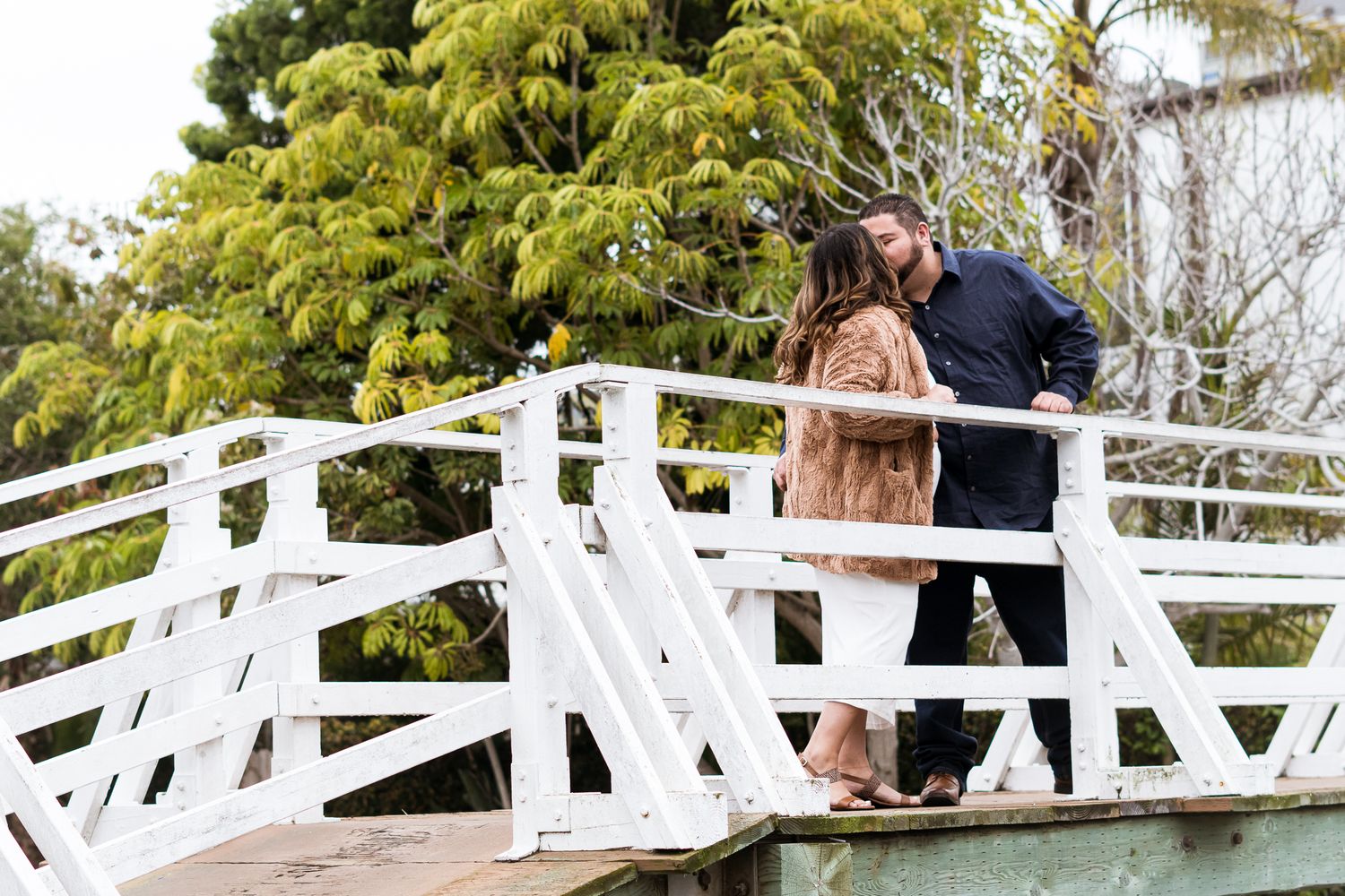 An engaged couple stands on a white wooden bridge in a garden setting during a romantic moment in Venice Beach CA.