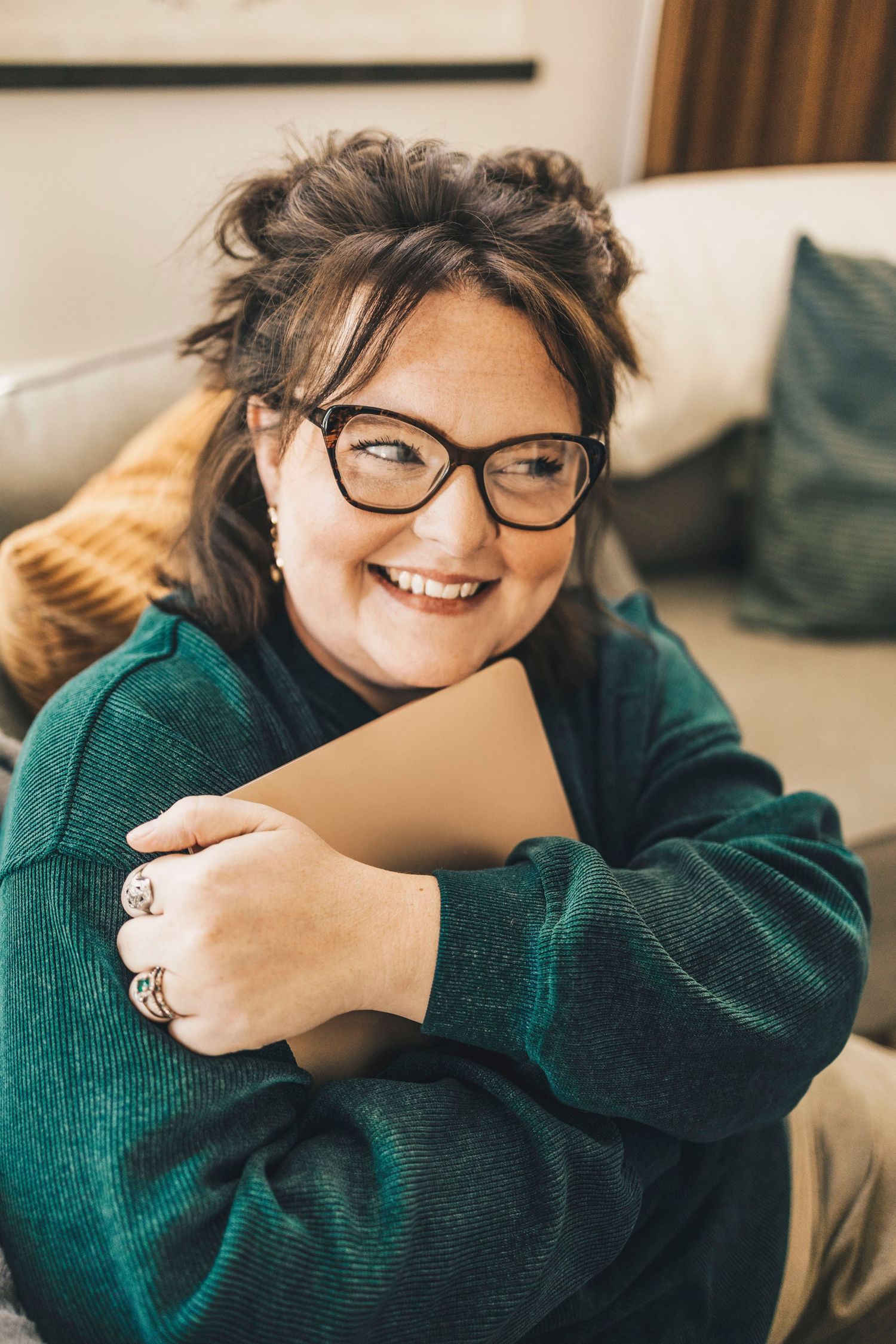 Smiling woman with glasses in green sweater hugging a laptop computer.