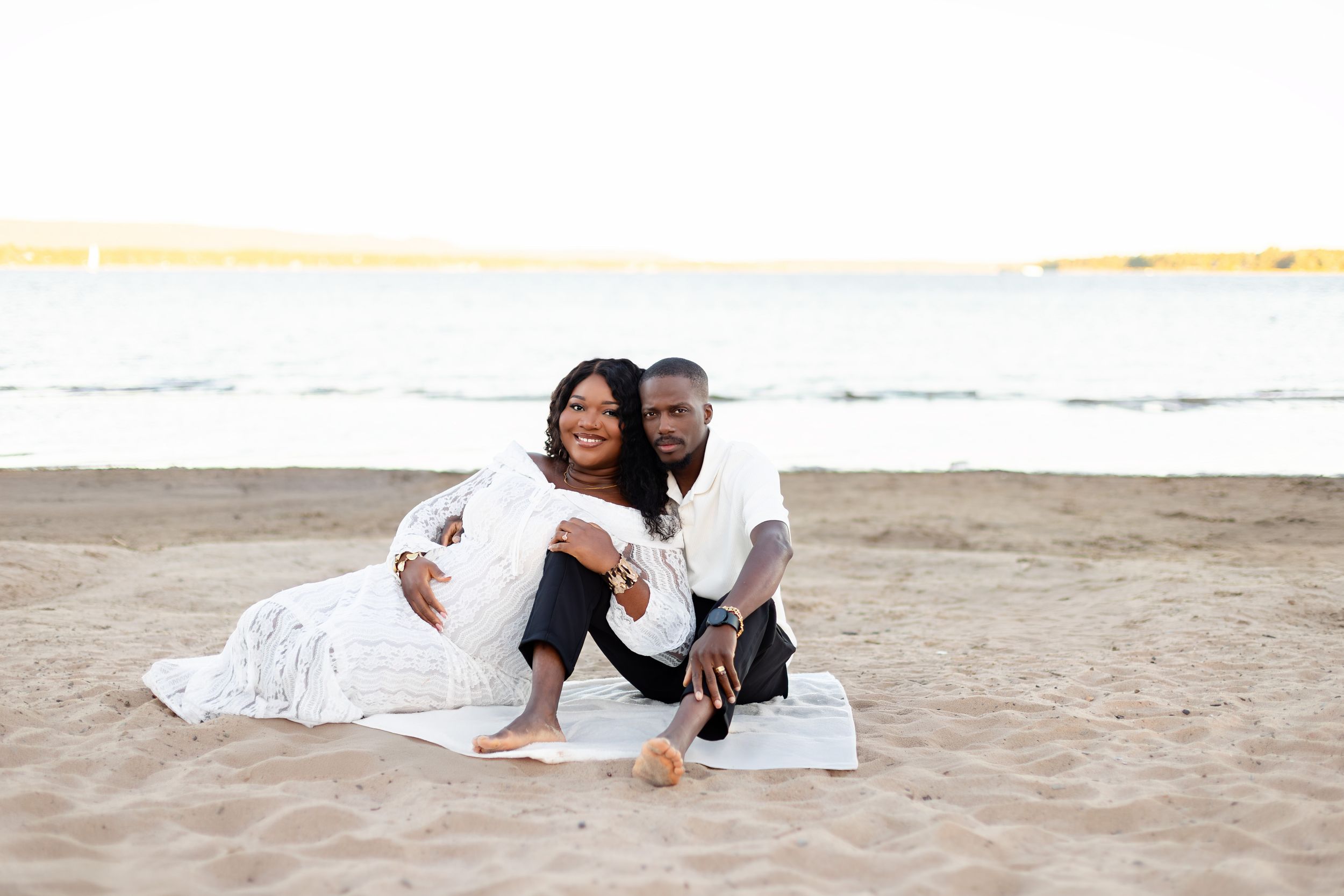 Happy couple in white outfits laying on beach sand during romantic photoshoot session.