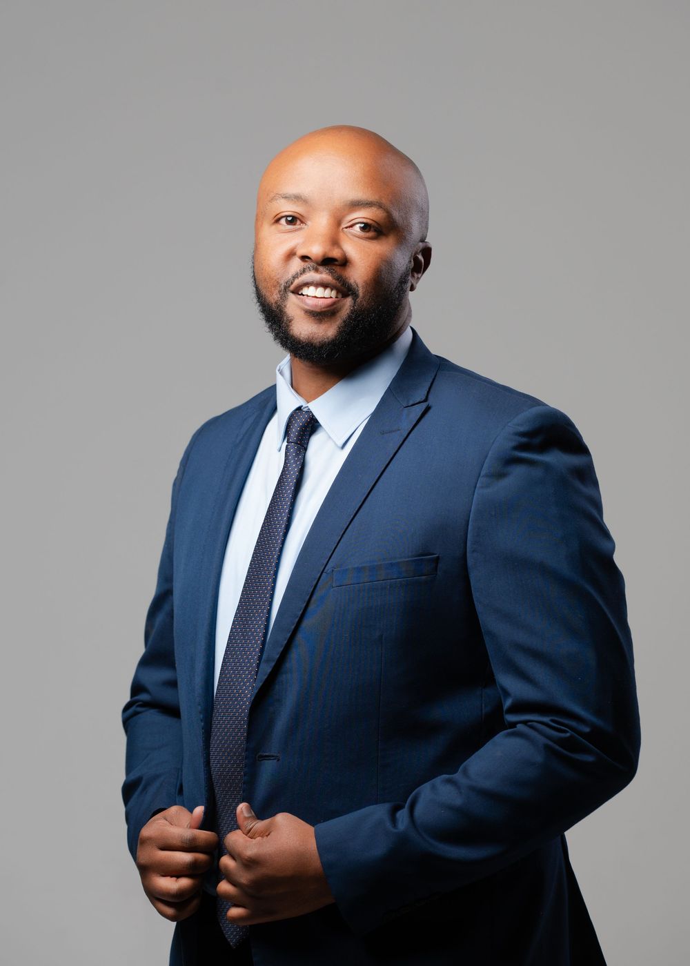Professional headshot of person in navy suit and tie against grey background.