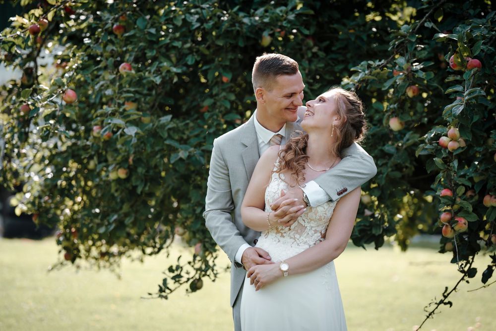 A couple shares a romantic kiss under lush green trees in the garden during their outdoor wedding celebration.