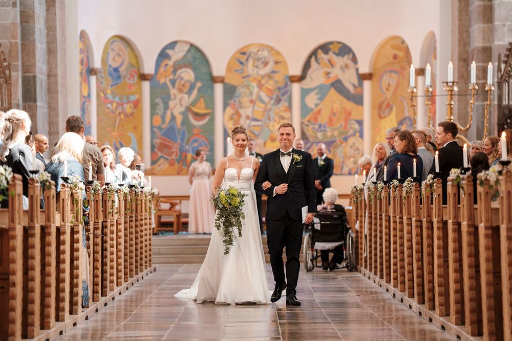 A couple walks down the aisle of a beautiful church with ornate religious murals and wooden pews.