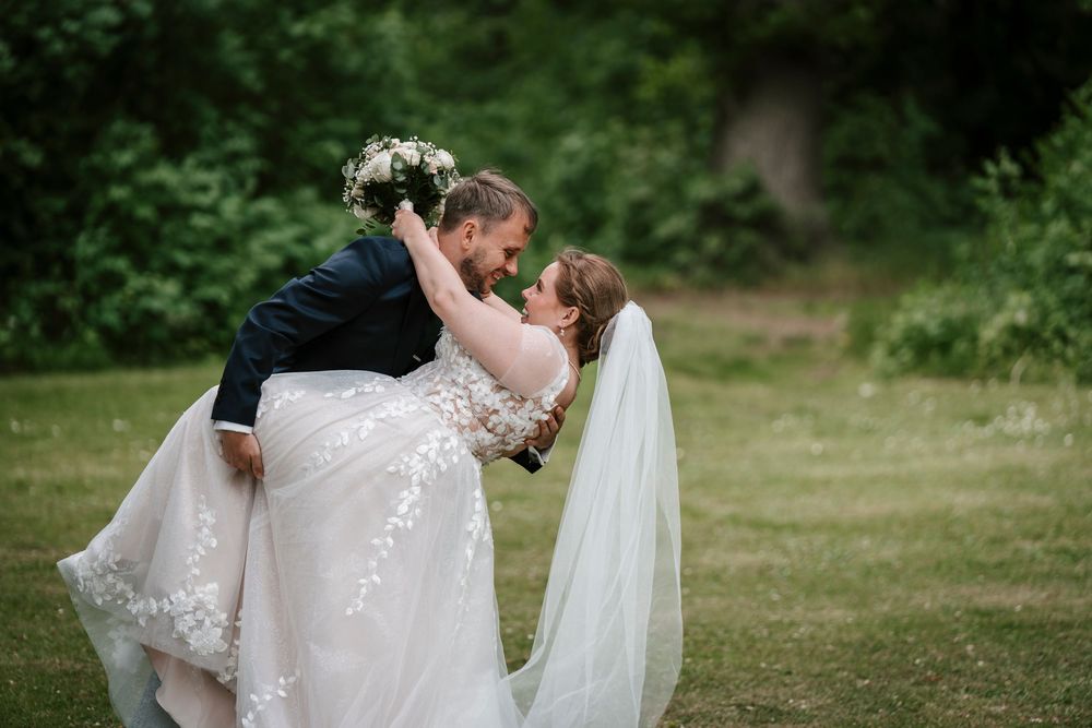 Groom lifts bride in lace wedding gown and veil, sharing a romantic moment outdoors on green grass.
