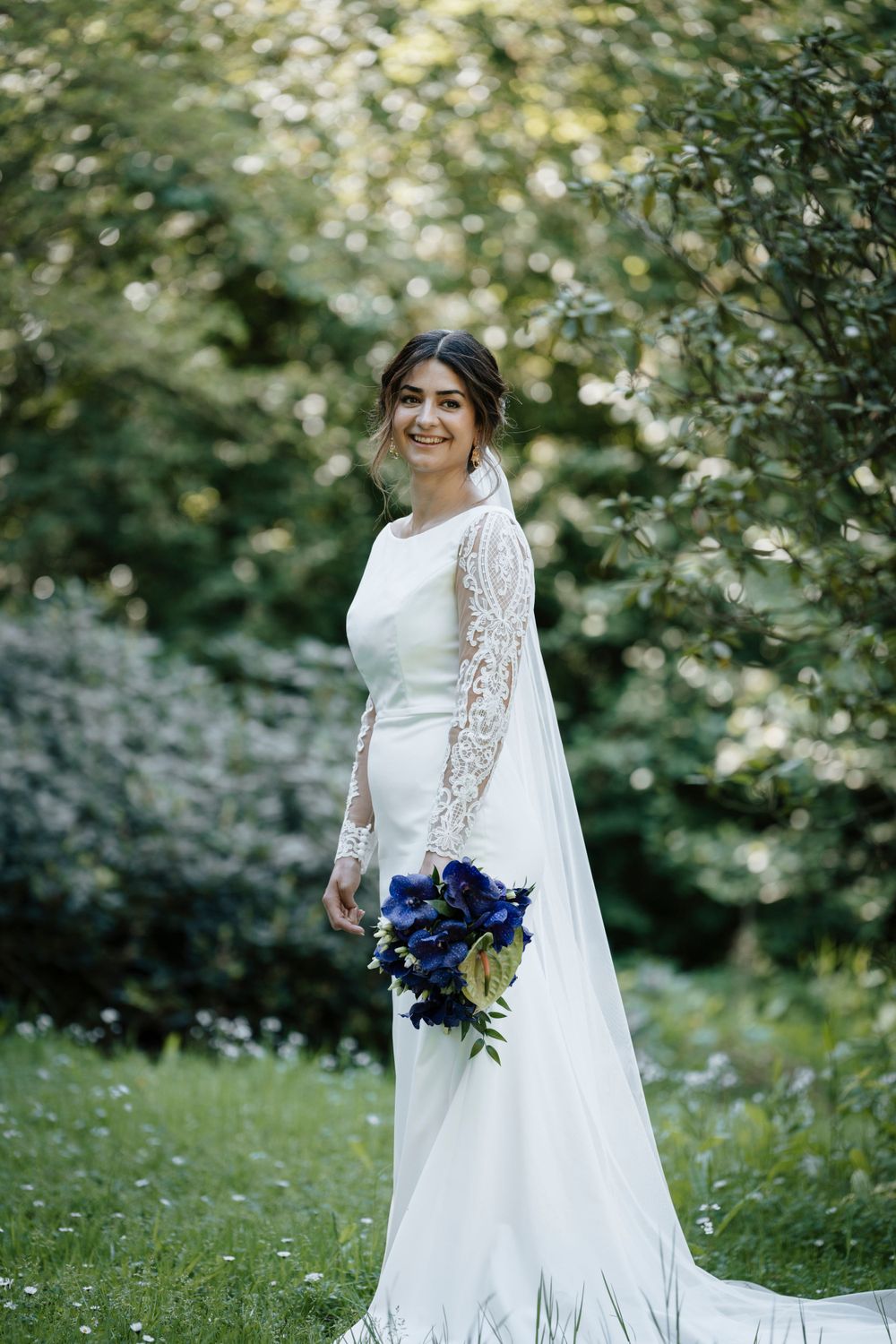 Brud i hvid kjole med blondeærmer, som holder sin brudebuket med blå blomster i  en park.