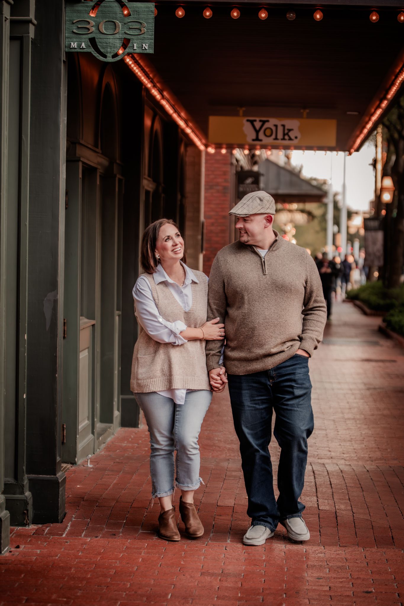 Couple walking together on a brick sidewalk holding hands outside storefronts at dusk.