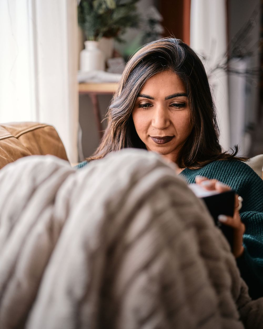 Person relaxes on cozy couch at home wearing green sweater with houseplants and white curtains in background.