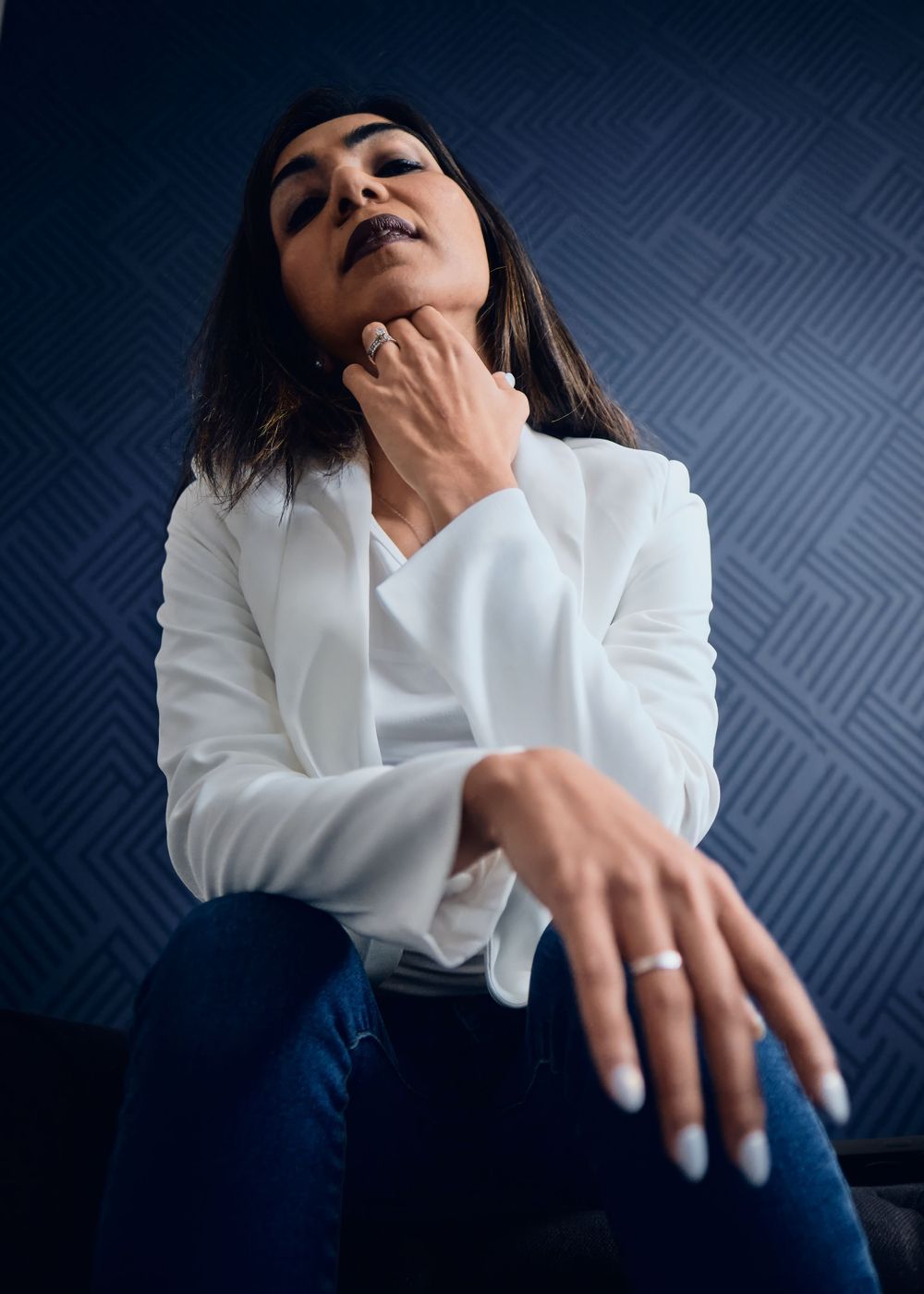 Editorial portrait of a model in a white long sleeve top and jeans posing dramatically against a dark blue patterned wall.