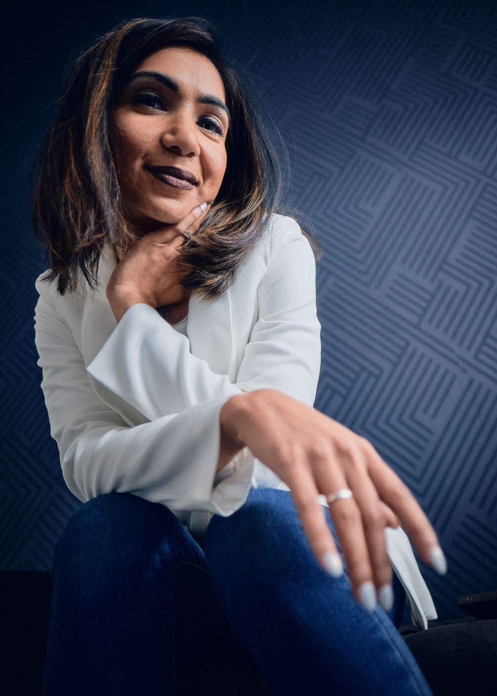 Editorial portrait of a model in a white blazer and jeans sitting casually against a textured blue wall.