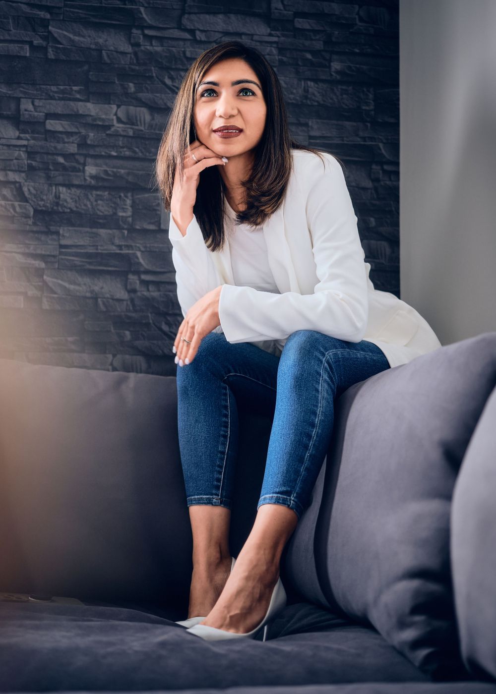 A thoughtful-looking woman in a white sweater and jeans sits relaxed on a gray couch against a textured dark wall.