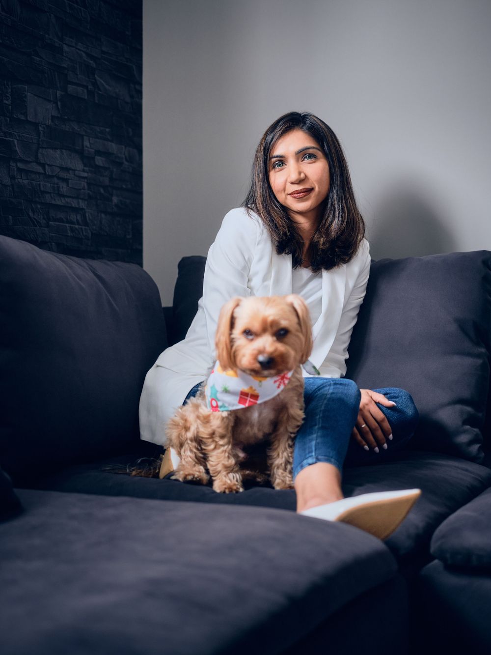 A woman relaxes on a dark gray couch with an adorable Yorkshire Terrier wearing a colorful collar.