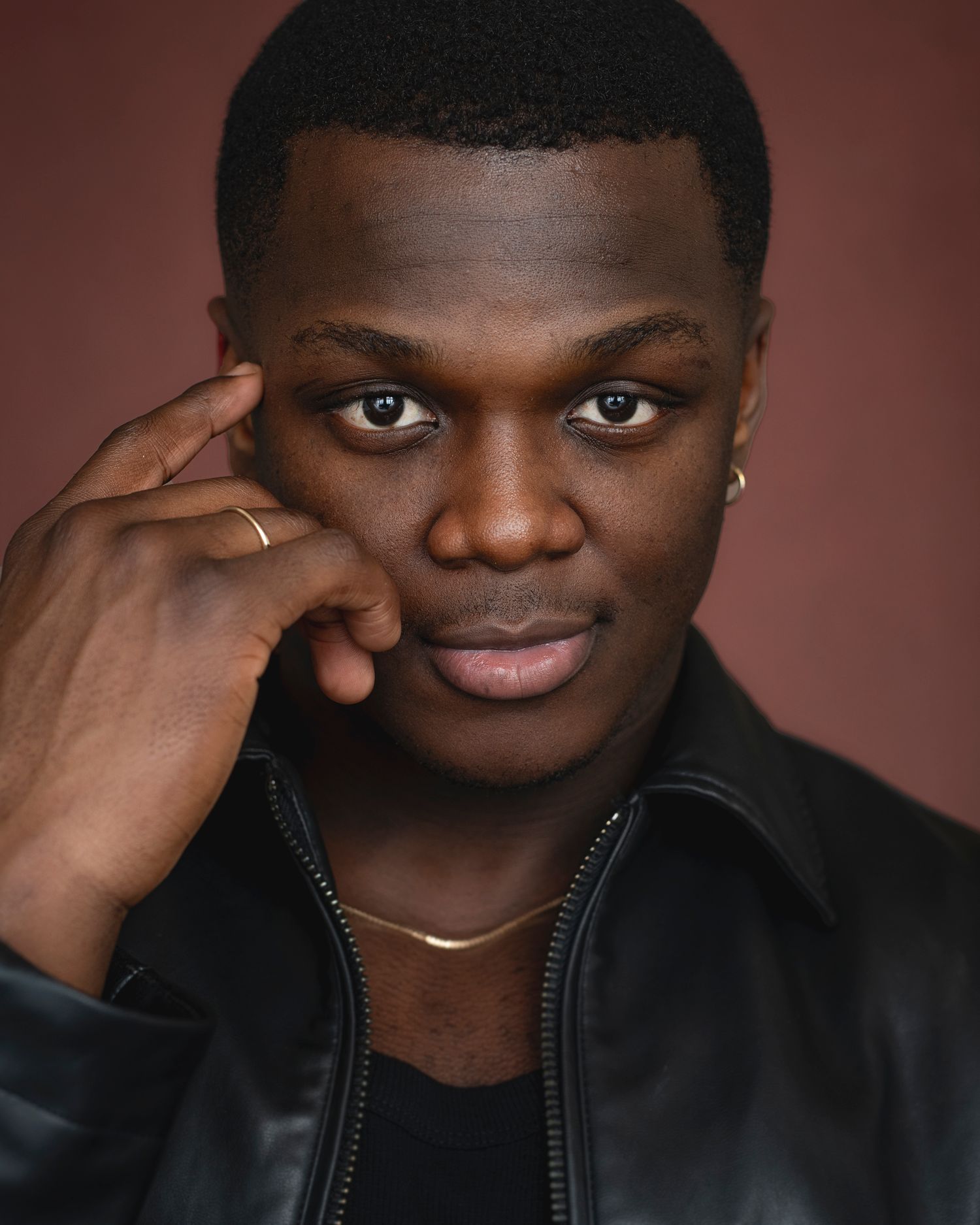 Close-up portrait with hand gesture wearing black jacket and gold necklace.