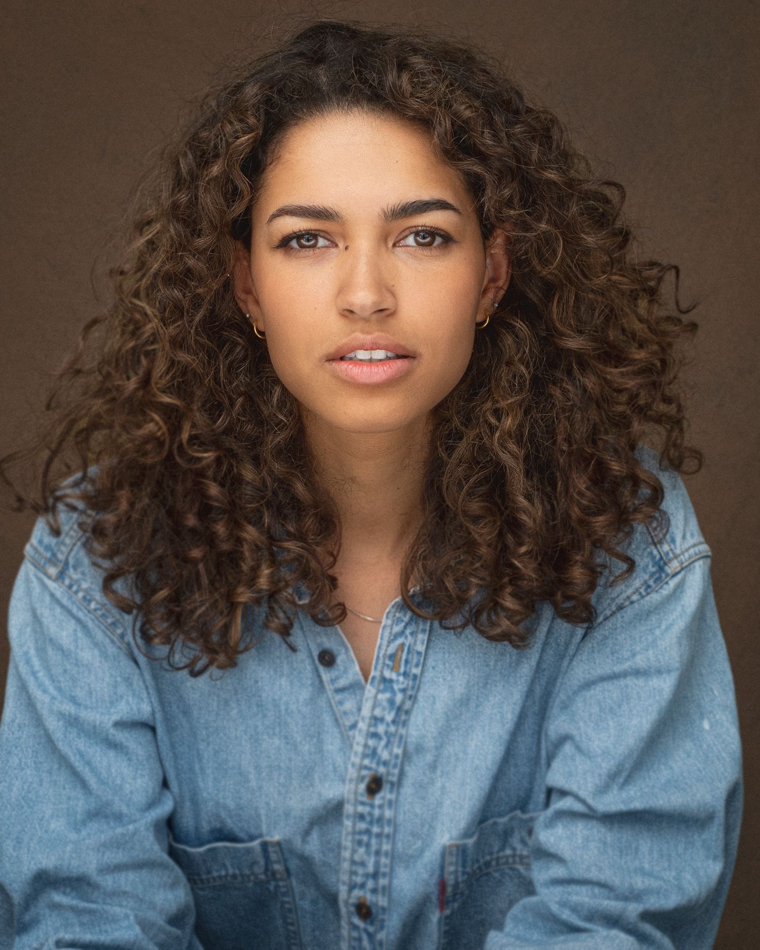 Studio portrait of person with natural curly hair wearing denim jacket against brown background.