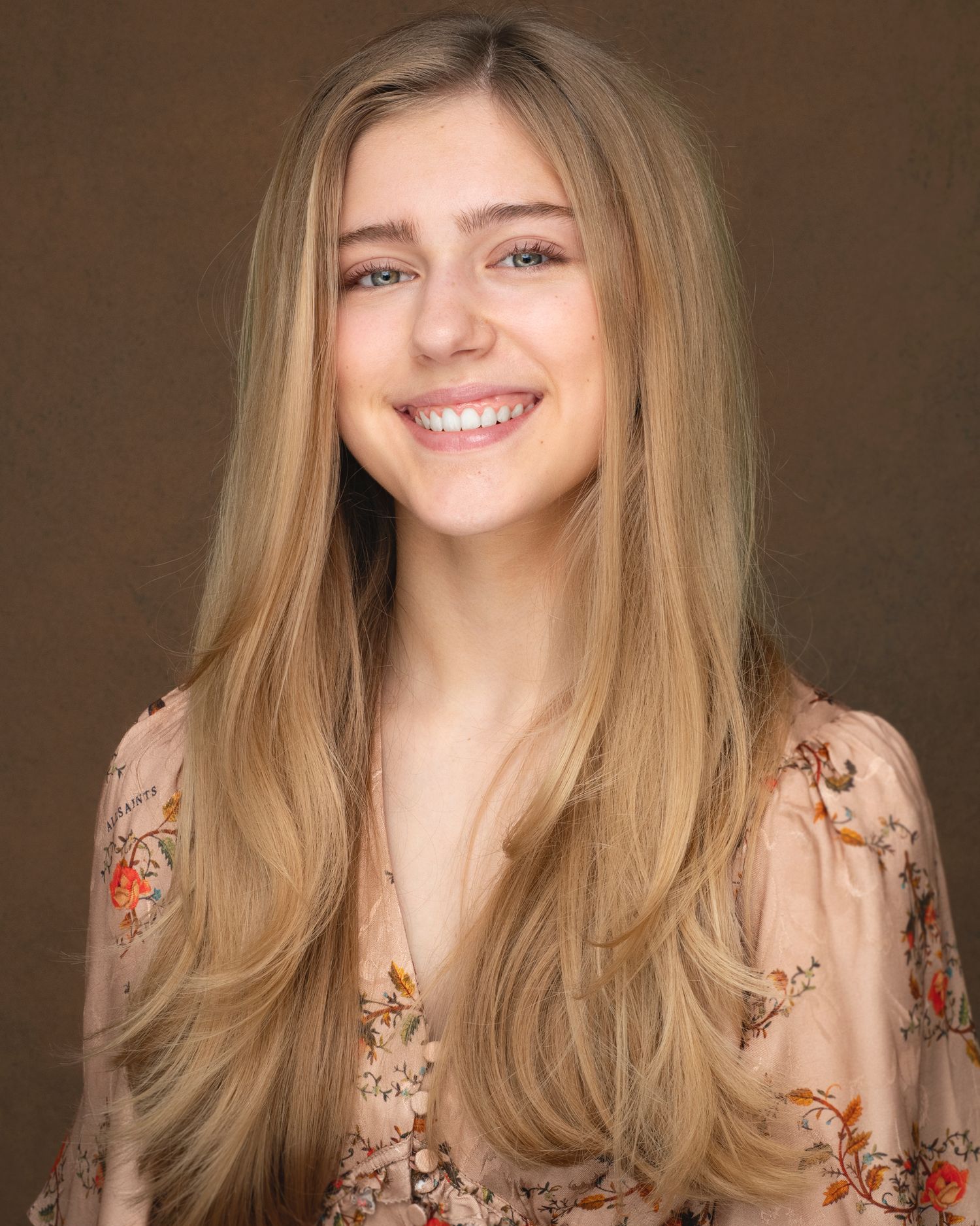 Portrait against brown backdrop of person with long blonde hair wearing floral print blouse.