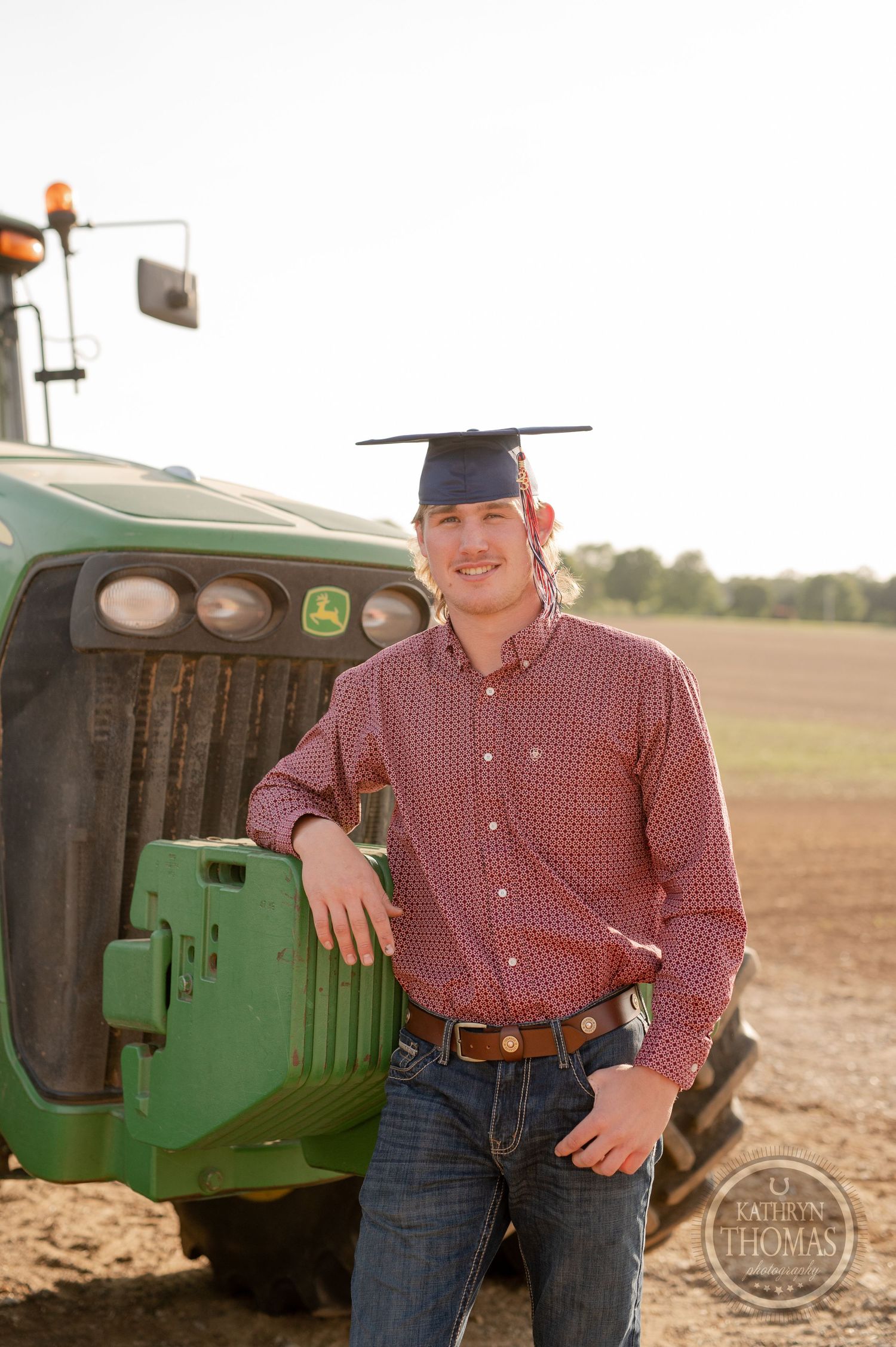 Person in graduation cap and red shirt poses with John Deere tractor in field.