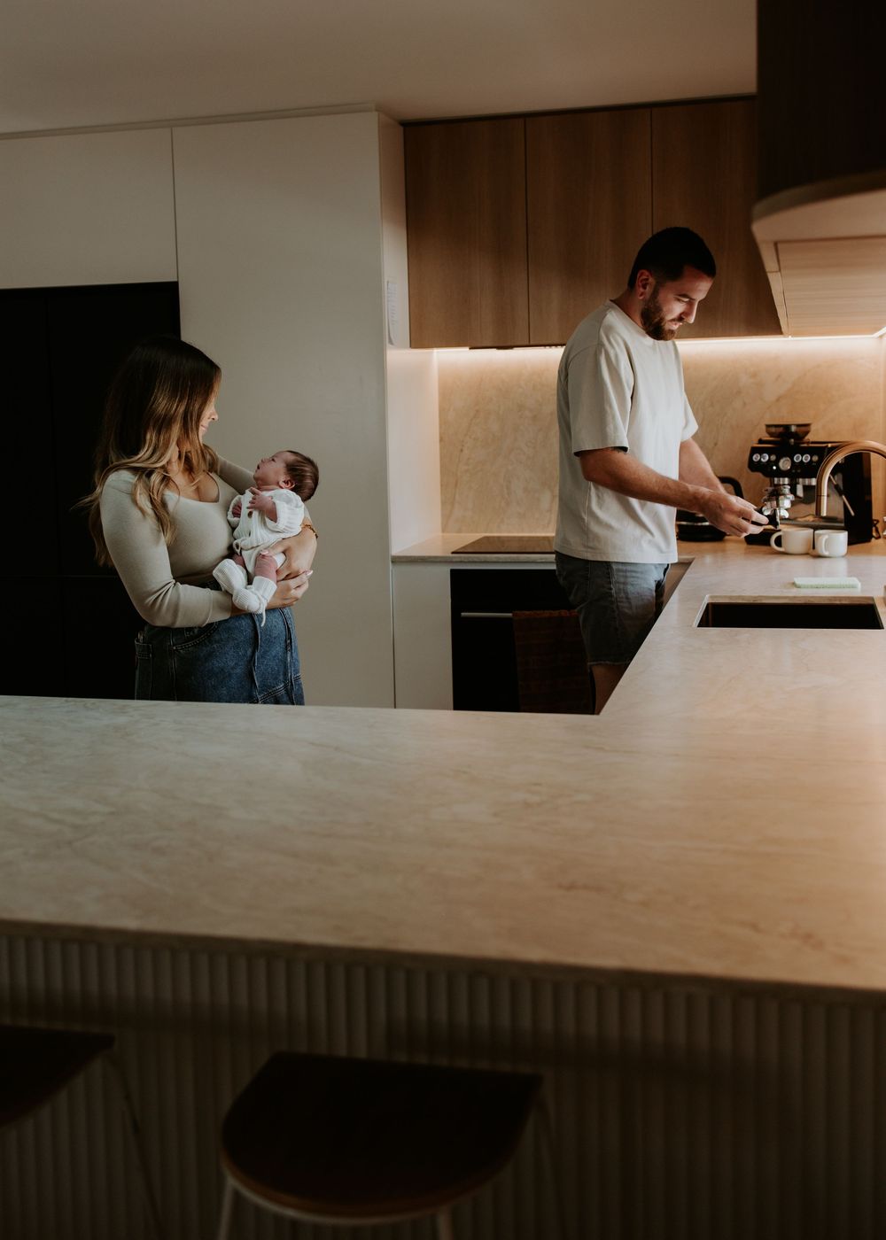A family spends time together in their modern kitchen as one parent holds a baby while the other makes coffee.