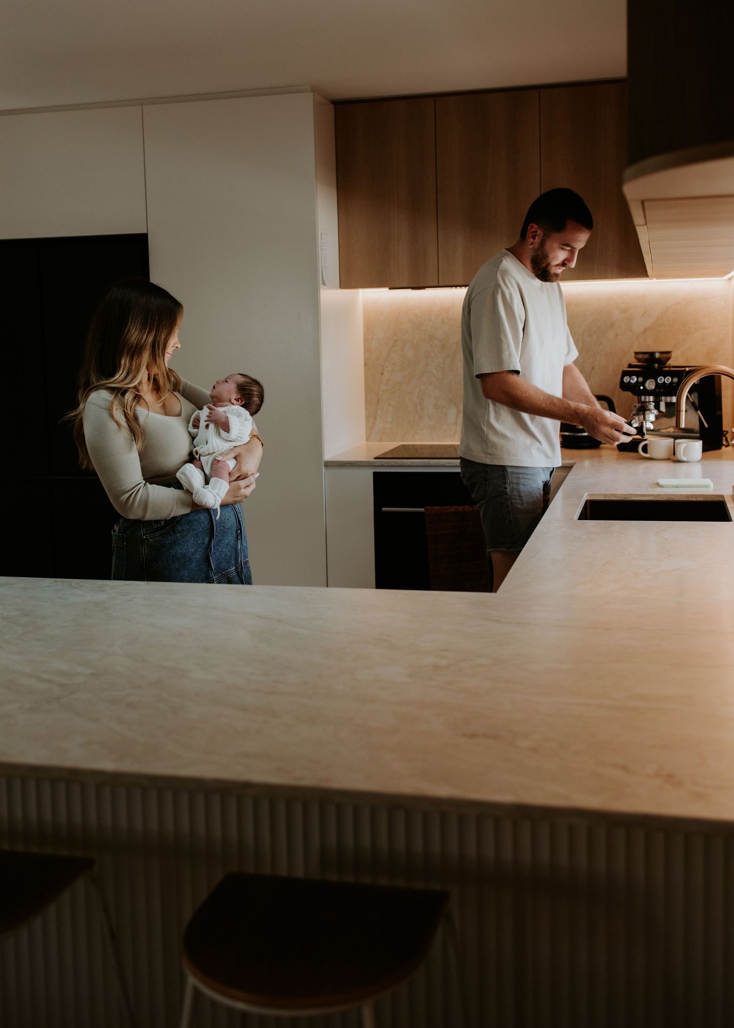 A family spends time together in their modern kitchen as one parent holds a baby while the other makes coffee.