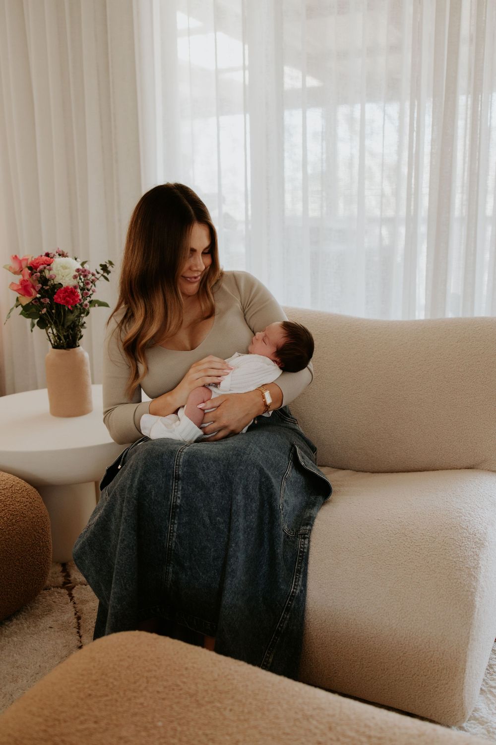 Mother and newborn baby share tender moments on beige couch in bright sunlit room with sheer curtains and flowers.