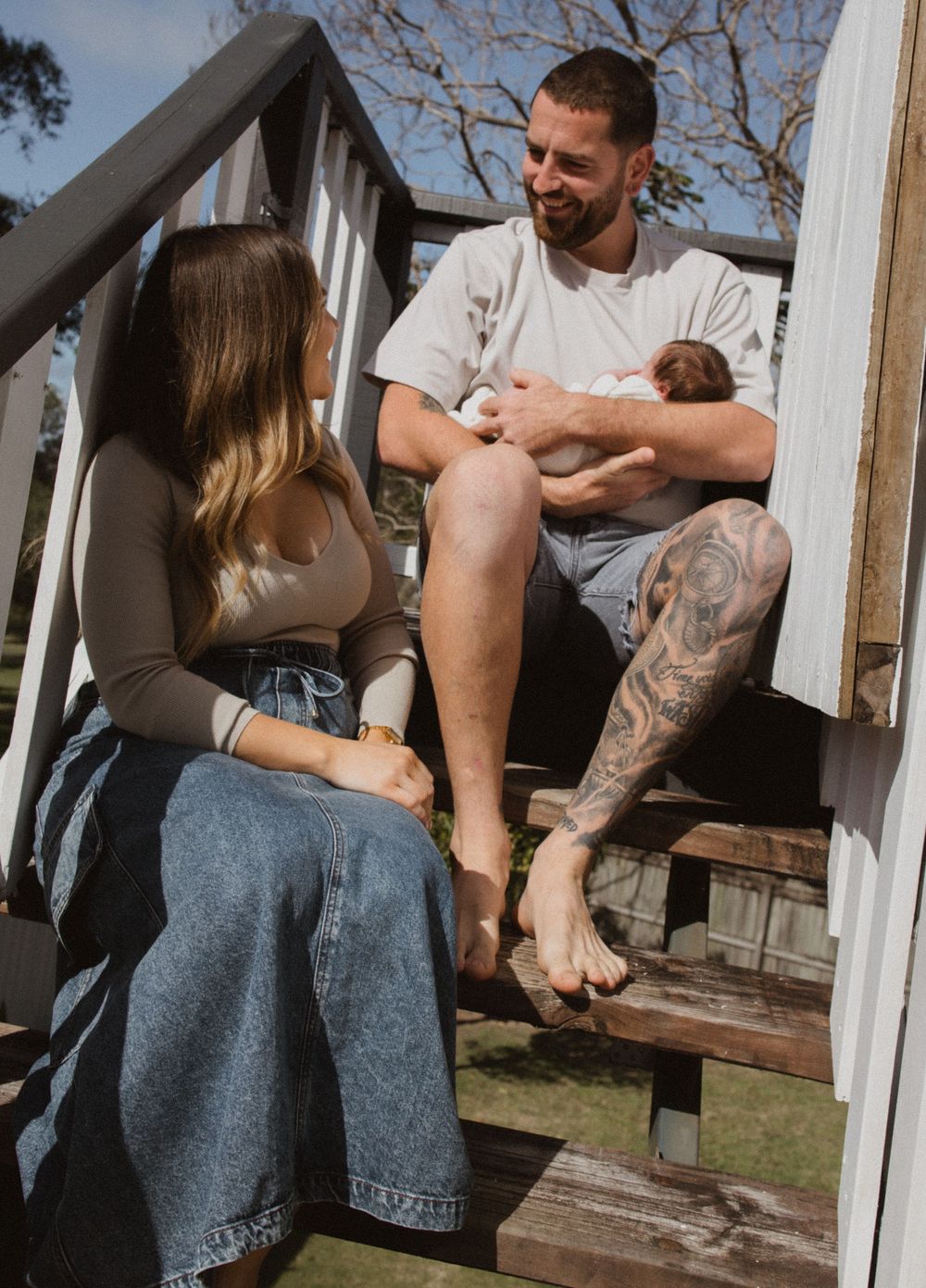 A couple sits on wooden steps outdoors while holding a newborn baby, with the man displaying a leg tattoo.
