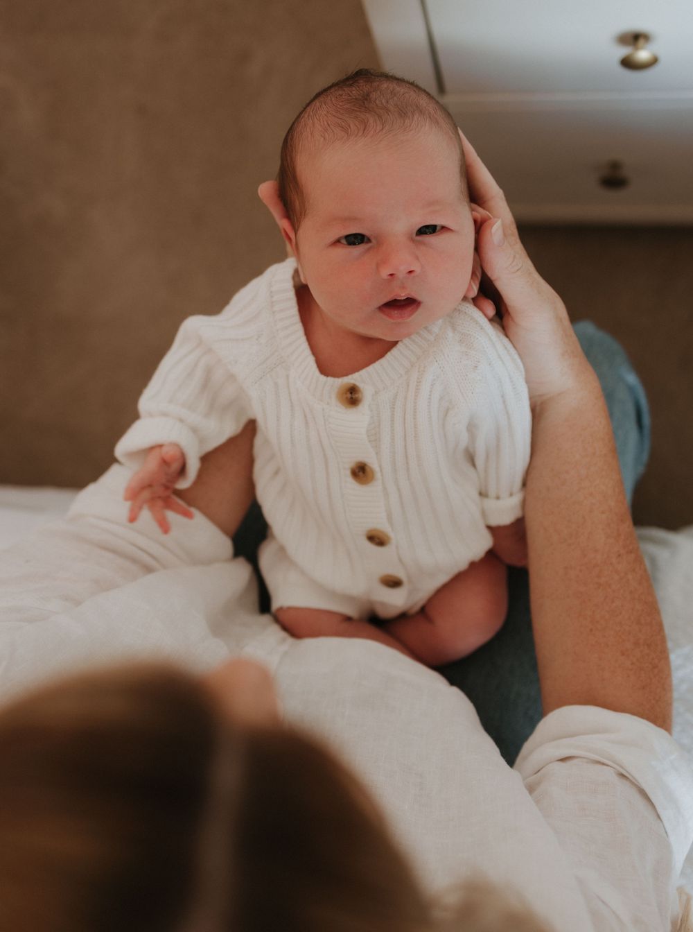 Baby wearing white button-up outfit lies comfortably on someone's lap in a warm, cozy indoor setting.