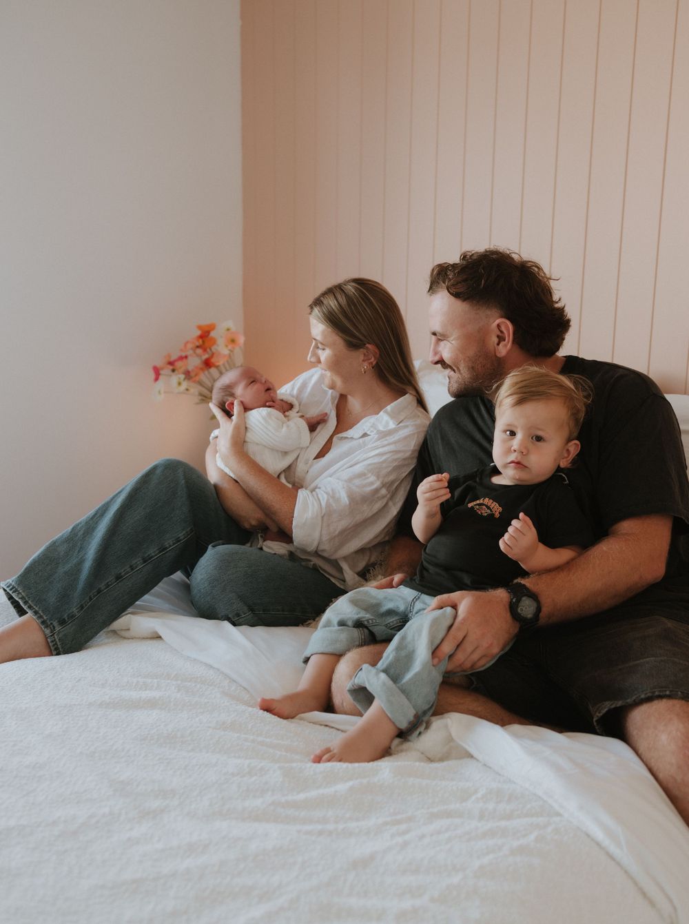 A family relaxes together on a white bed while watching something on a tablet device.