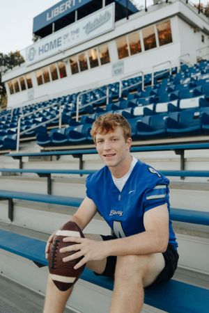 High school senior in his football gear, sitting in stands at high school with football.