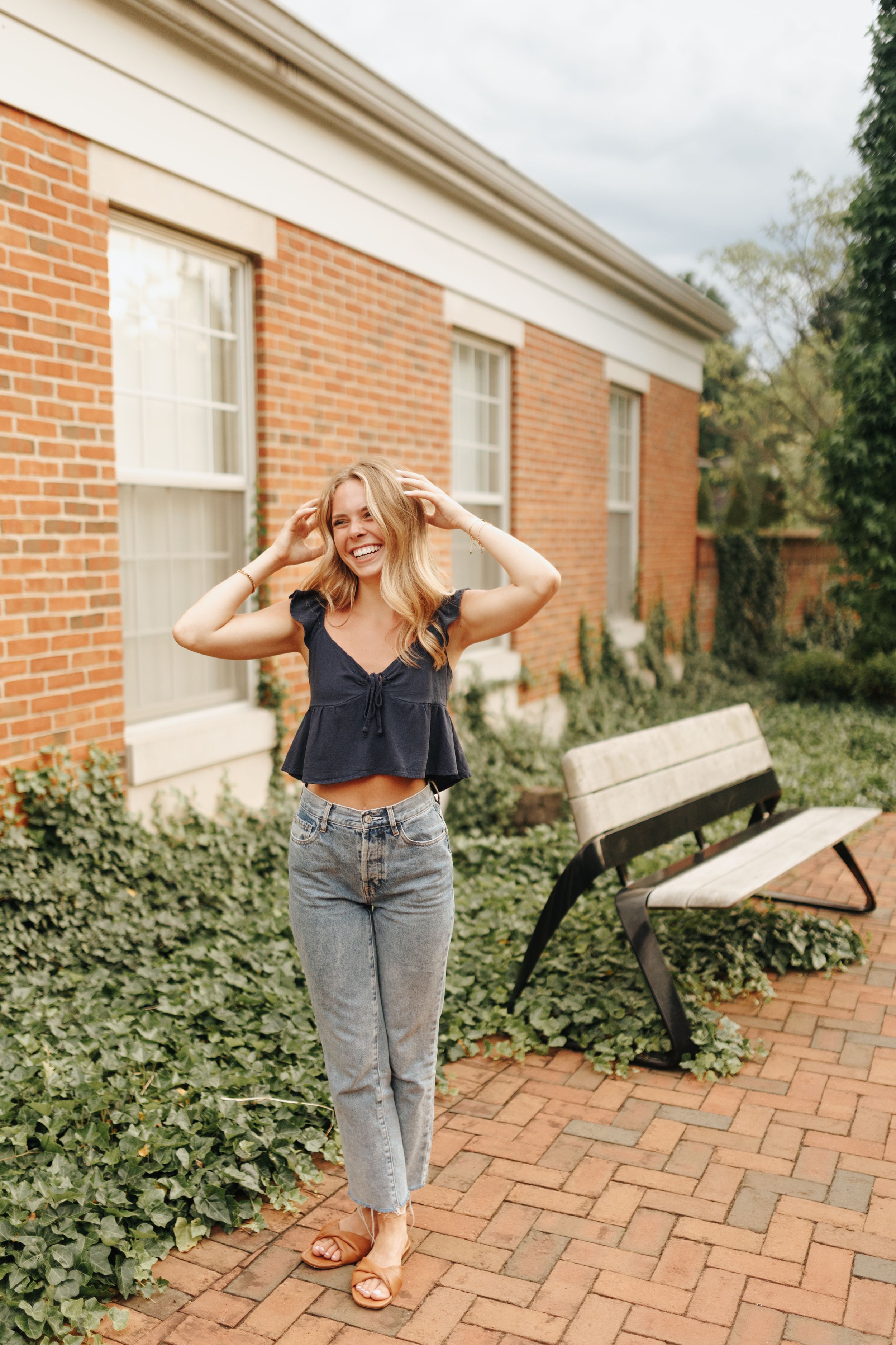 Senior posing with a big smile outside a charming brick building, wearing a stylish outfit.
