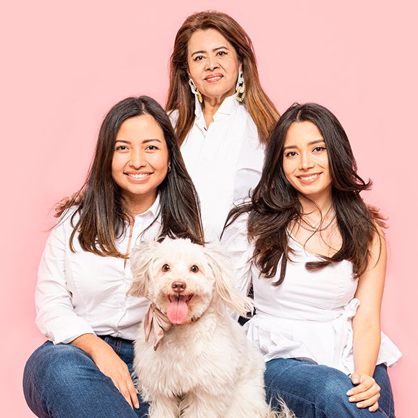 Three people in white tops and jeans pose with a fluffy white dog against a pink backdrop.