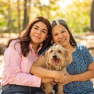 A heartwarming outdoor portrait showing two people and their small fluffy dog against a sunlit forest backdrop.