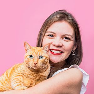 A happy orange tabby cat being held against a pink background.