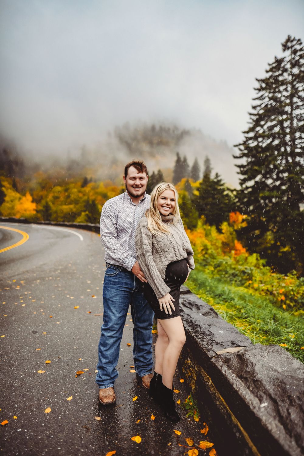 A couple poses playfully on a misty mountain road surrounded by fall foliage.